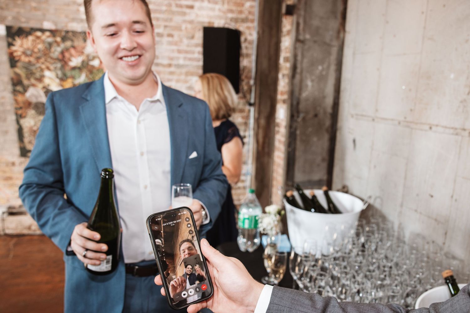 Smiling man in blue blazer holds champagne bottle while someone shows him a photo on a smartphone.