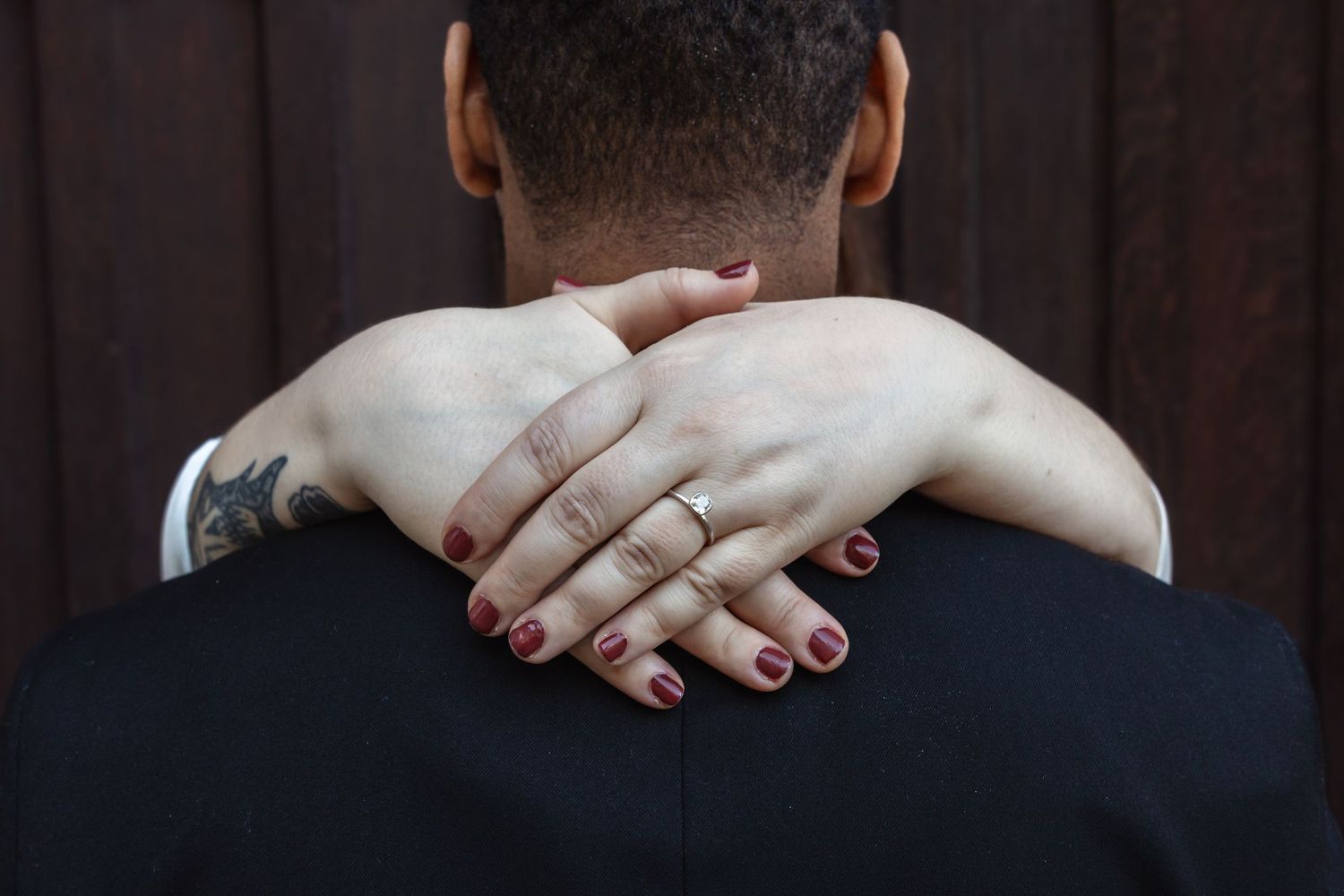 Bride embraces groom from behind showing wedding ring and red nails.