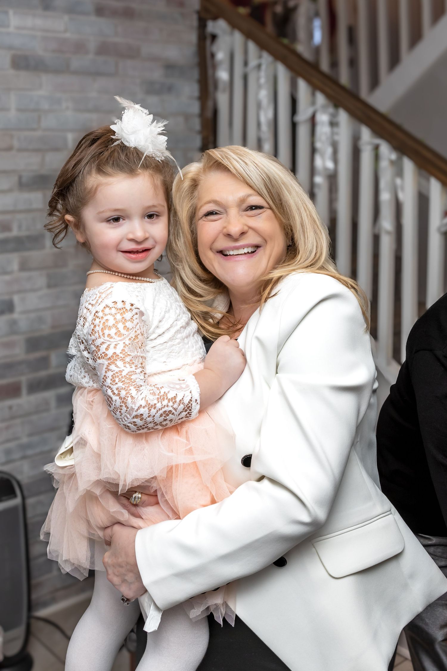 Smiling grandmother in white blazer holds young girl in pink tutu dress at wedding.