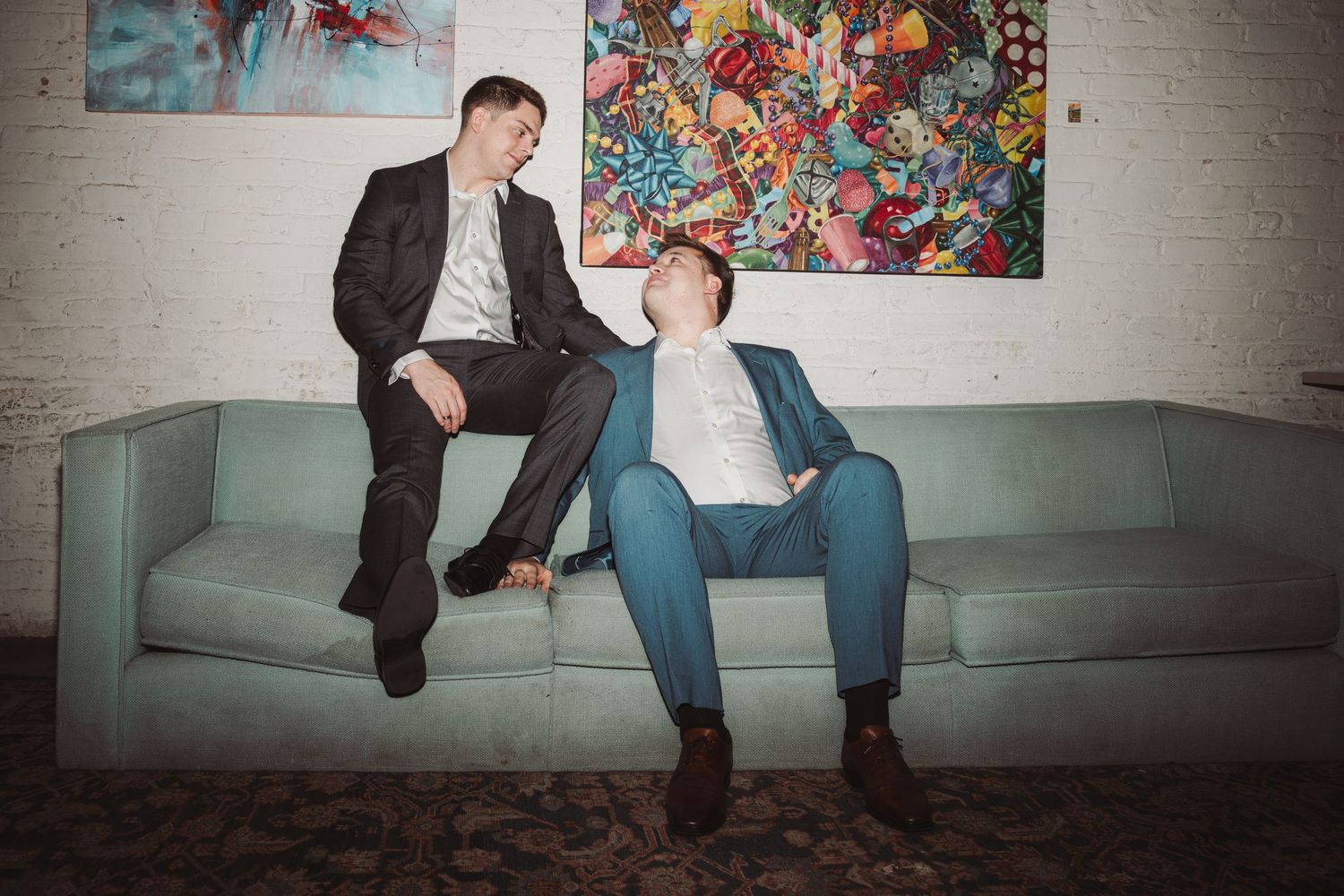 Two young men in suits relaxing on a mint green sofa with colorful artwork behind them.