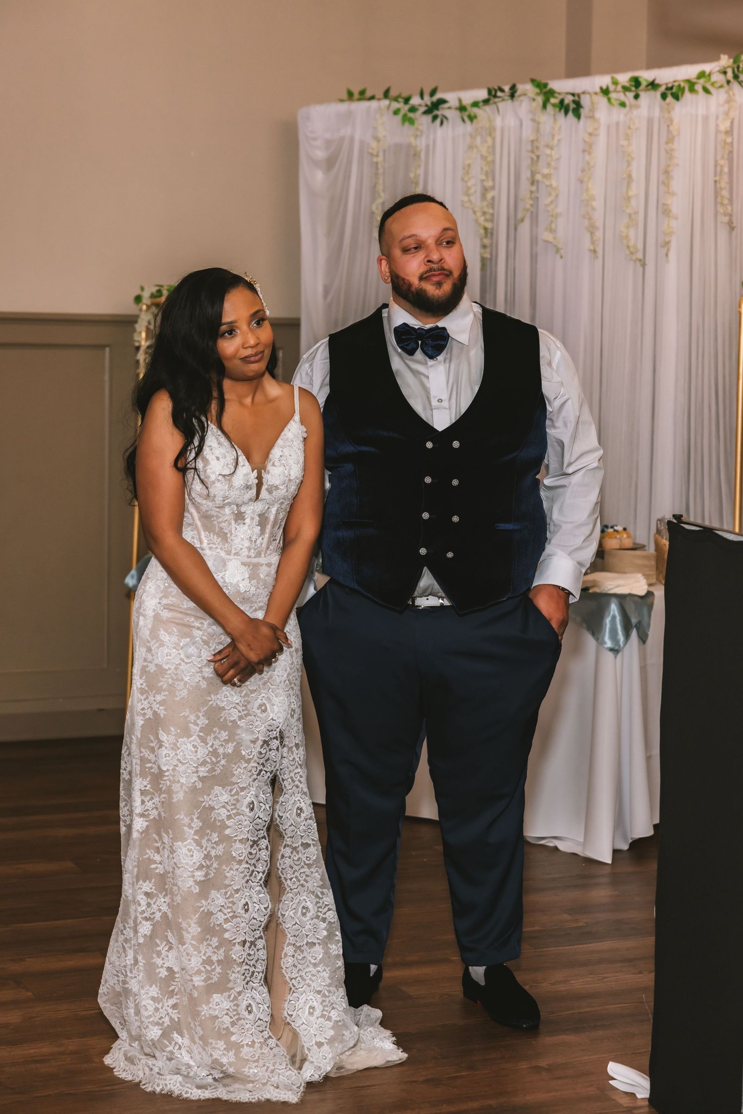 Bride in lace wedding gown and groom in navy vest and bow tie stand together at their wedding reception.