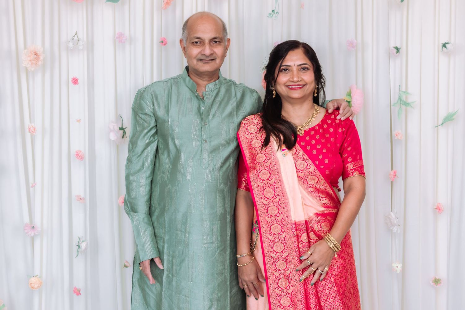 Indian couple posing together, man in green kurta and woman in pink saree, against floral backdrop.