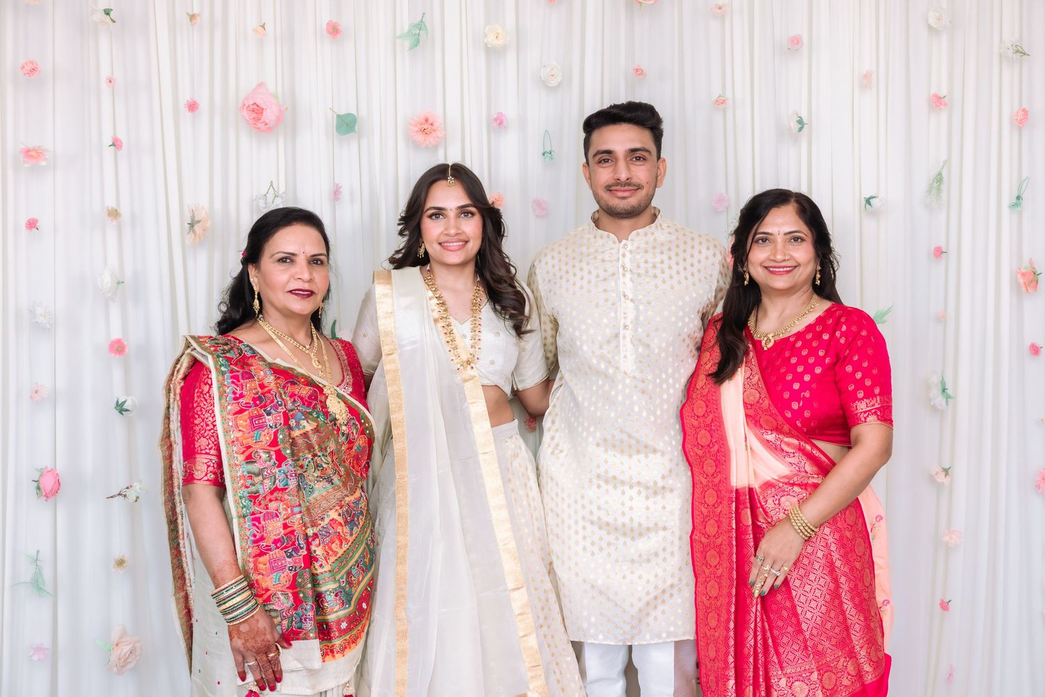 Four people in traditional Indian attire pose together at a wedding celebration with floral backdrop.
