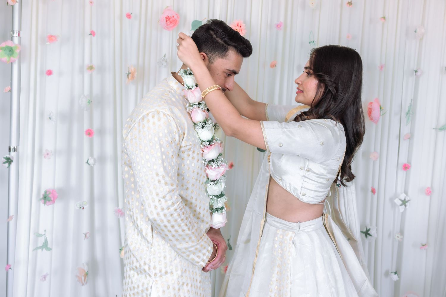 Bride placing floral garland on groom during traditional Indian wedding ceremony against white floral backdrop.