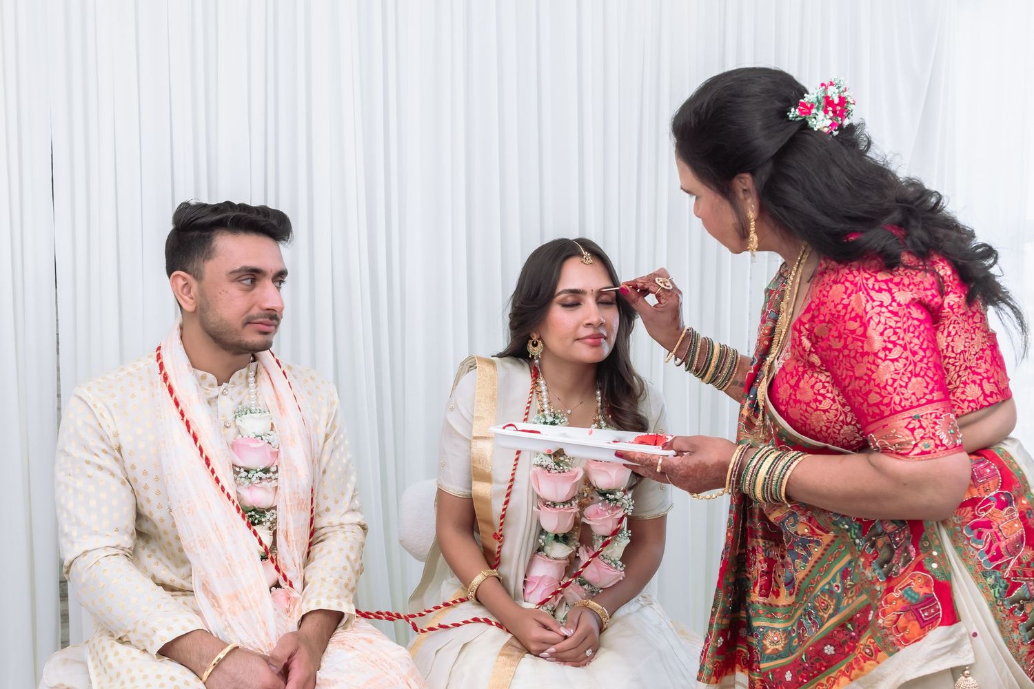 Elder woman performing traditional Indian wedding ritual with tikka plate for seated bride and groom.