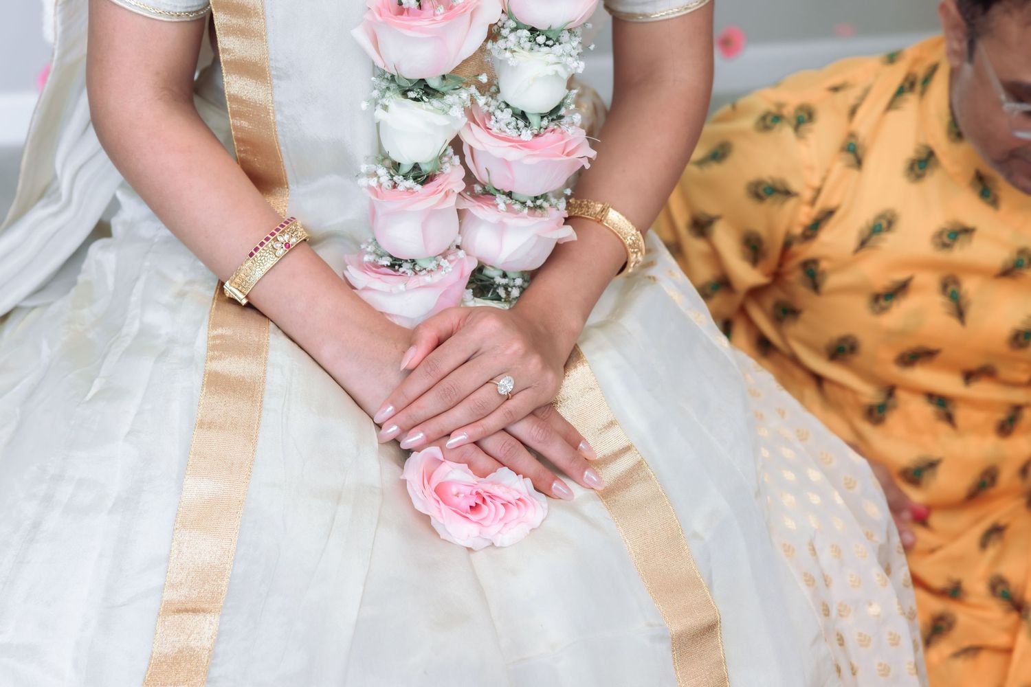 Bride holding pink rose garland in white and gold saree with engagement ring visible.