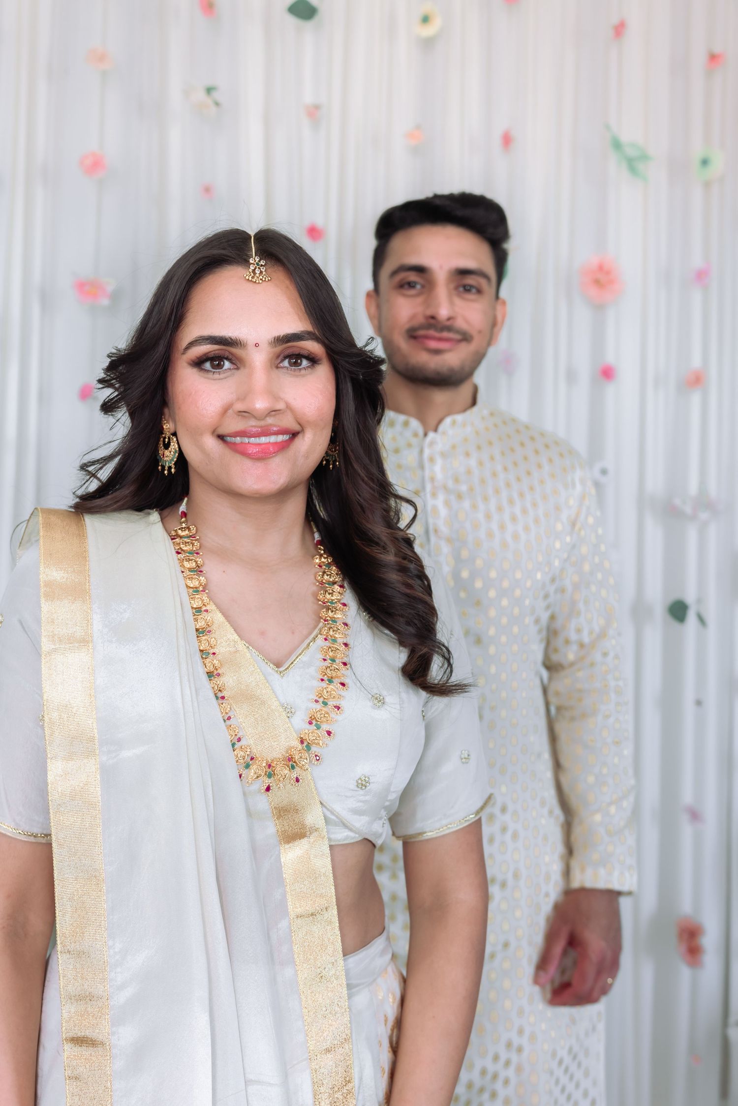 South Asian bride in white lehenga with gold jewelry smiling confidently with groom standing behind.