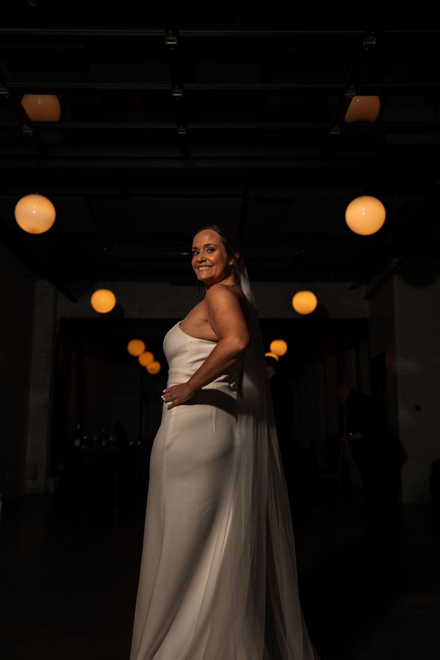 Bride in sleek white wedding gown poses dramatically in shadow with warm globe lights glowing in dark venue.