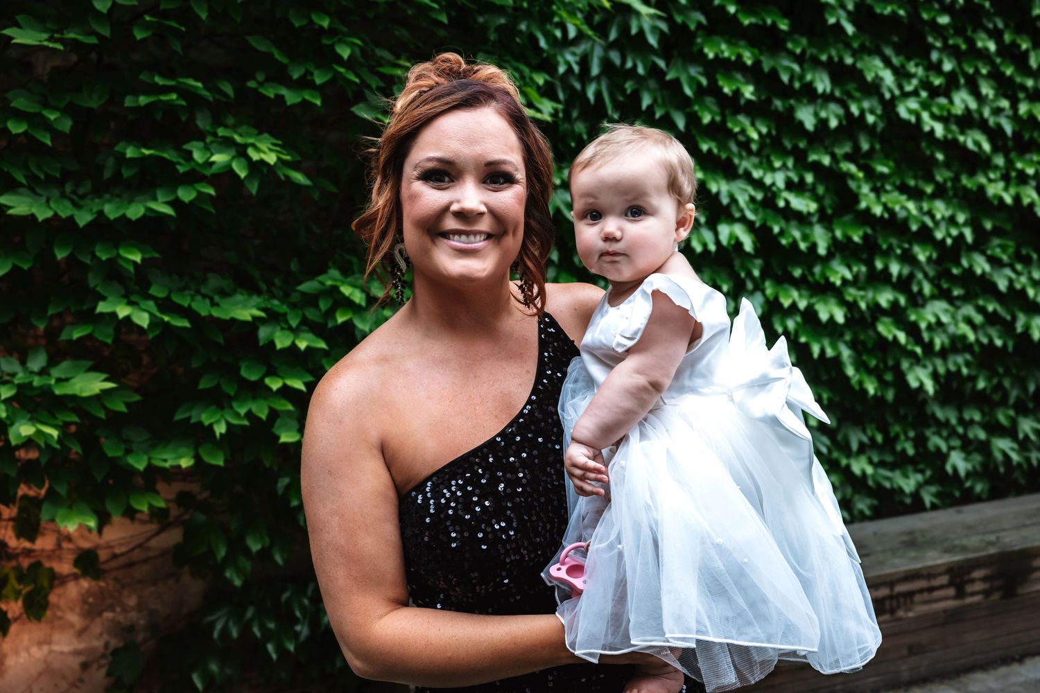Smiling woman in black dress holding baby girl in white dress before ivy wall.