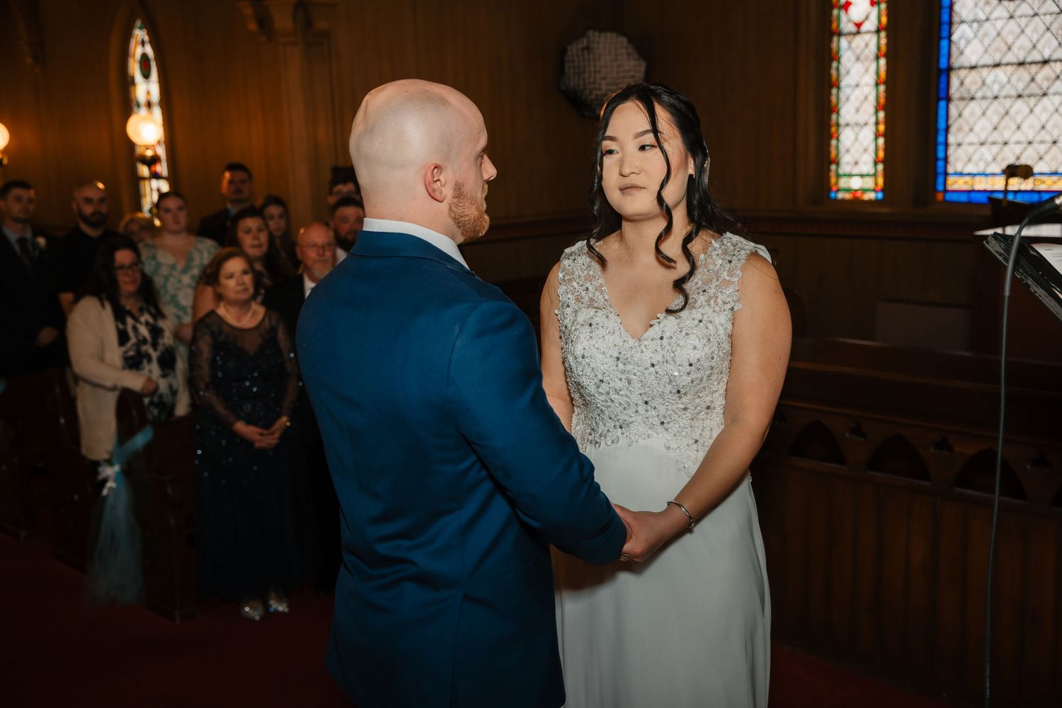 Bride and groom hold hands at wedding ceremony altar with guests seated in church pews behind them.