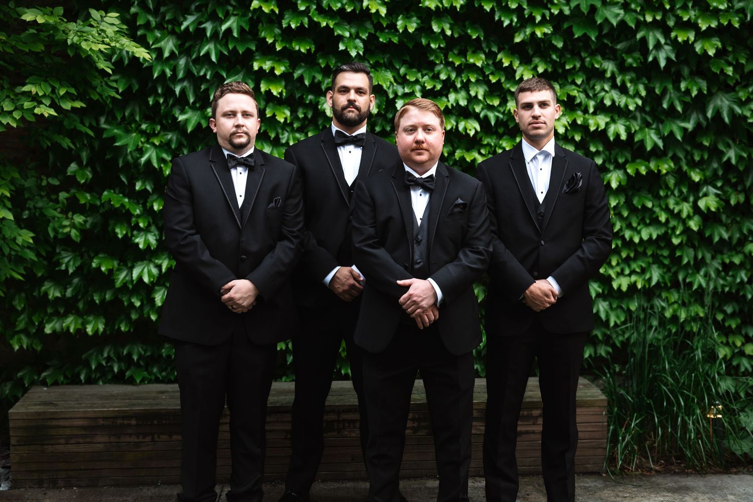 Four groomsmen in black tuxedos posing in front of a green ivy wall outdoors.