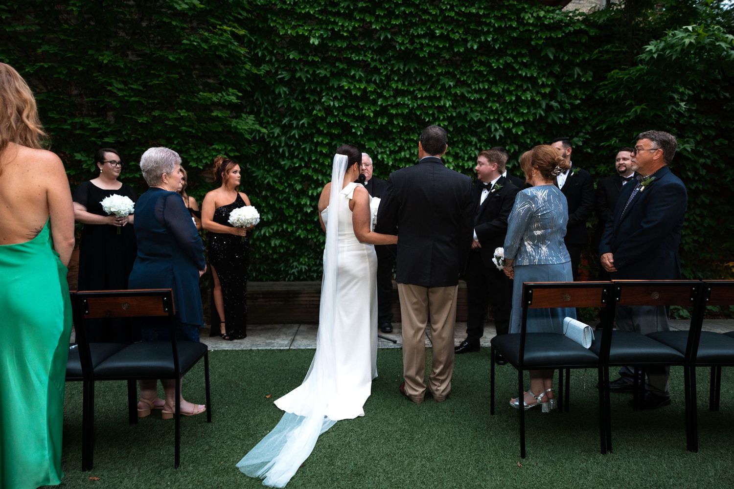 A bride in a white gown with veil approaches the altar in an outdoor garden wedding ceremony.