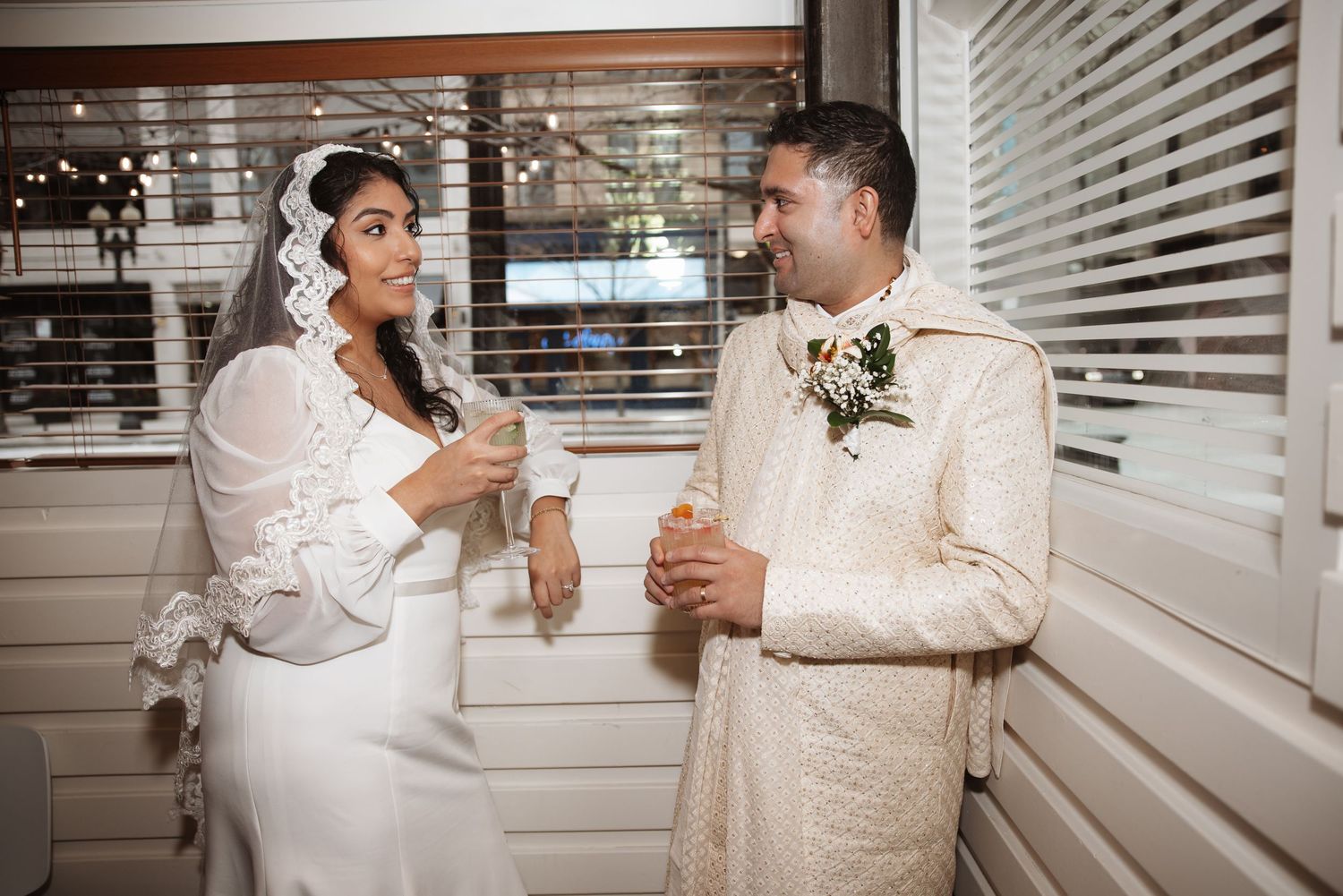 Bride in white gown with lace veil and groom in ivory sherwani toasting with cocktails near window blinds at wedding reception.