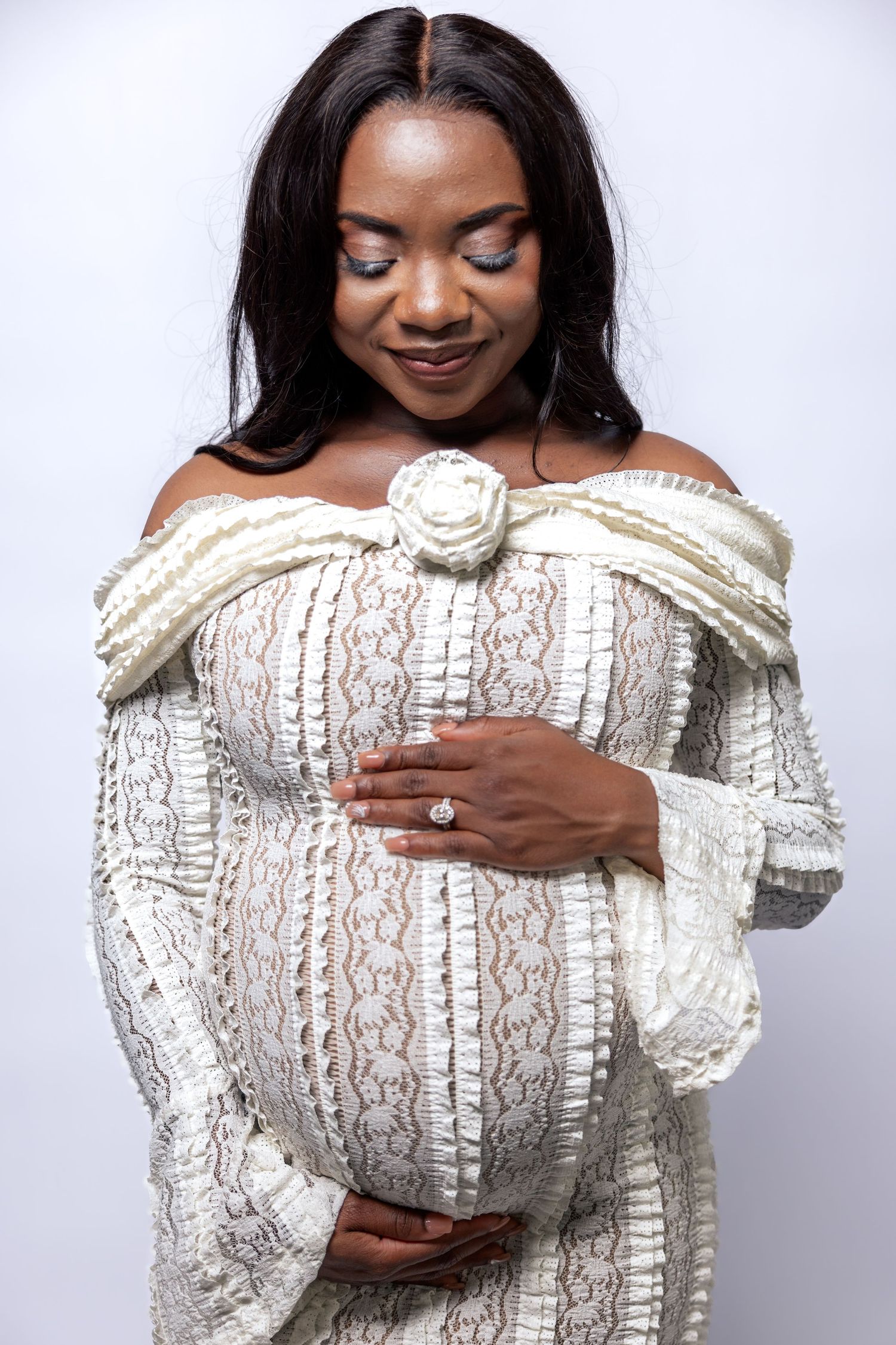 Pregnant Black woman in white lace off-shoulder gown looking down at baby bump, smiling.