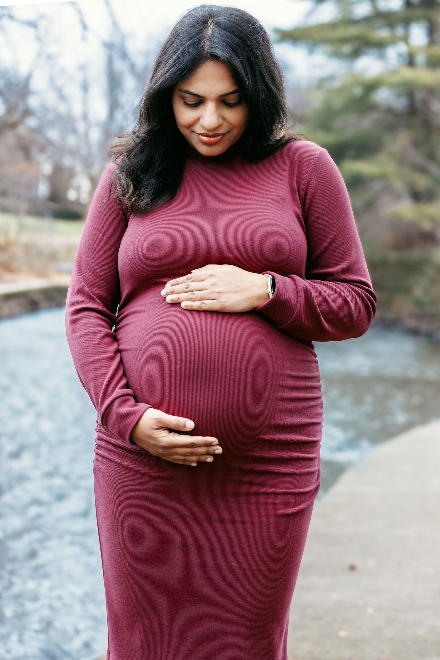 Smiling pregnant woman in burgundy dress gently holding her baby bump outdoors near a pond.