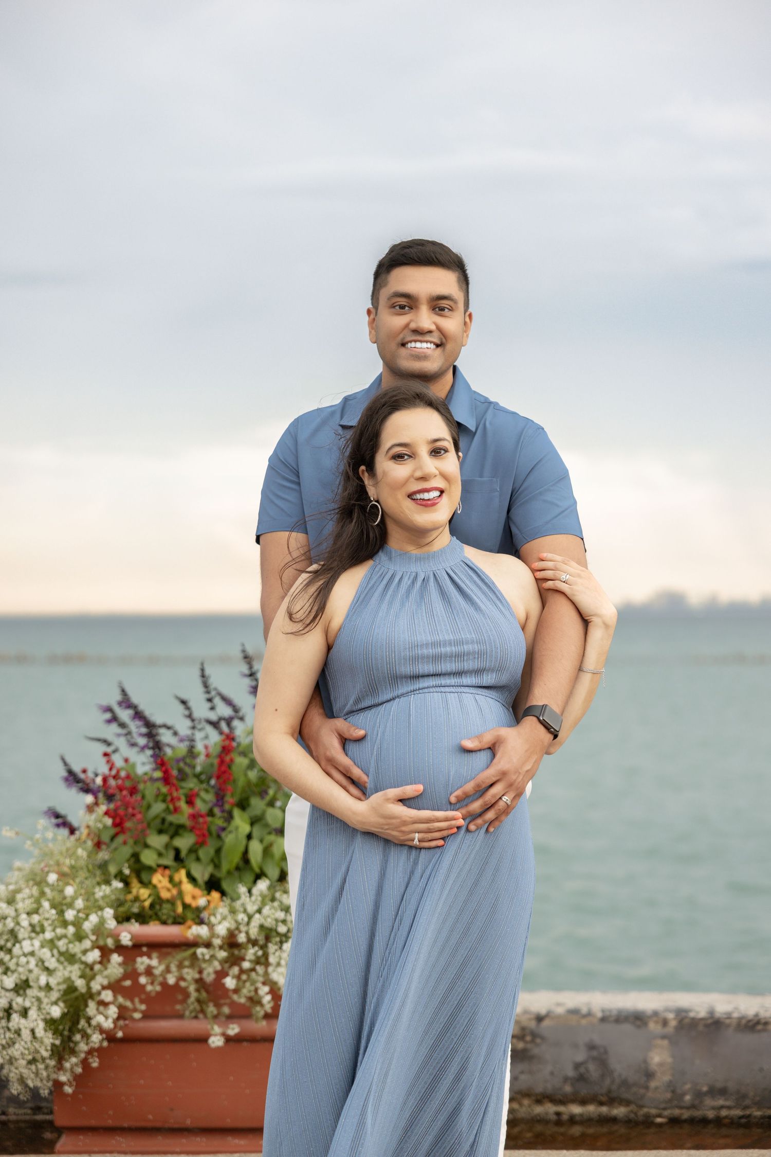 Smiling expectant couple posing by the lake, man embracing pregnant woman in blue halter dress, colorful flowers nearby.