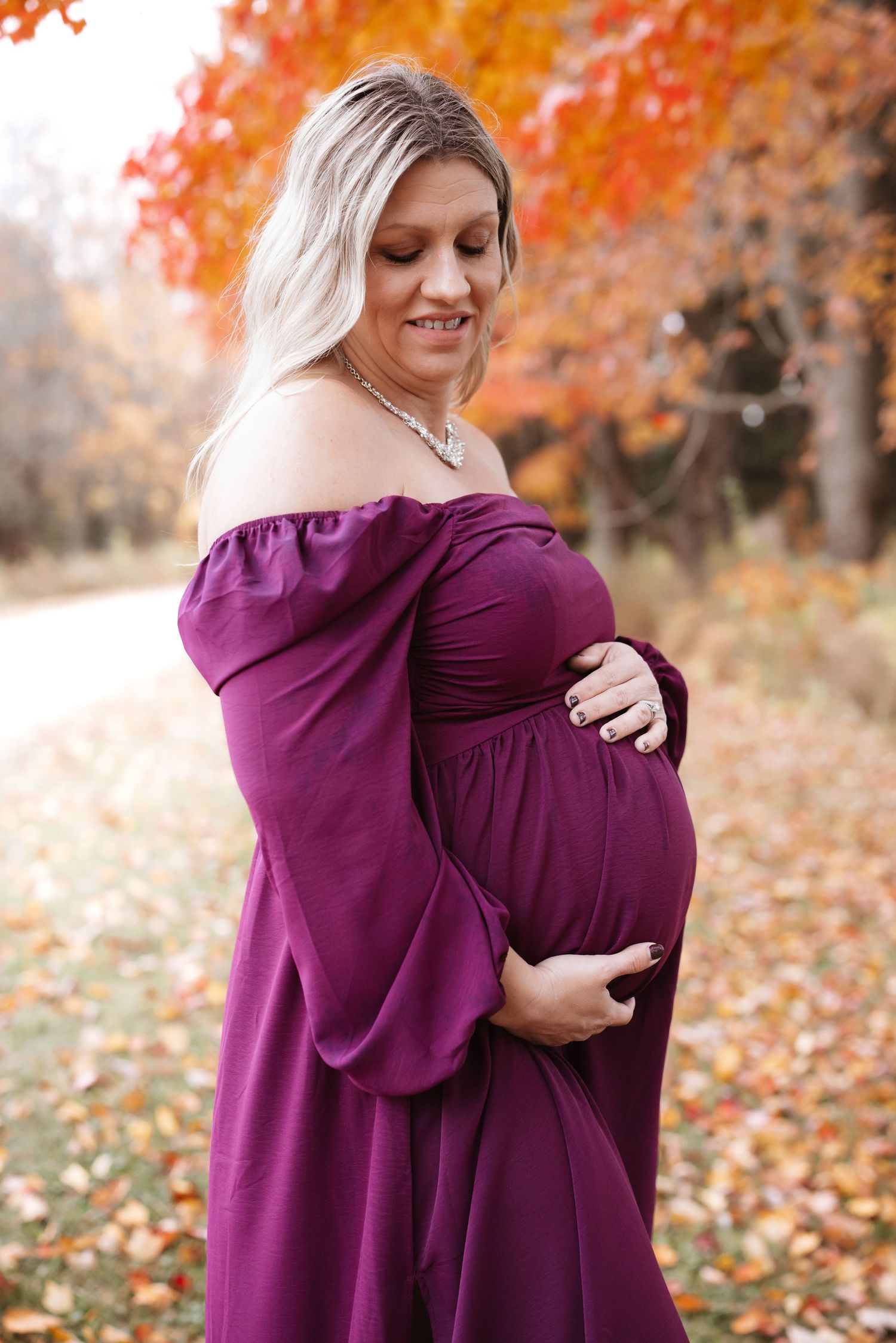 Pregnant woman in elegant purple off-shoulder gown tenderly holding baby bump in autumn setting.