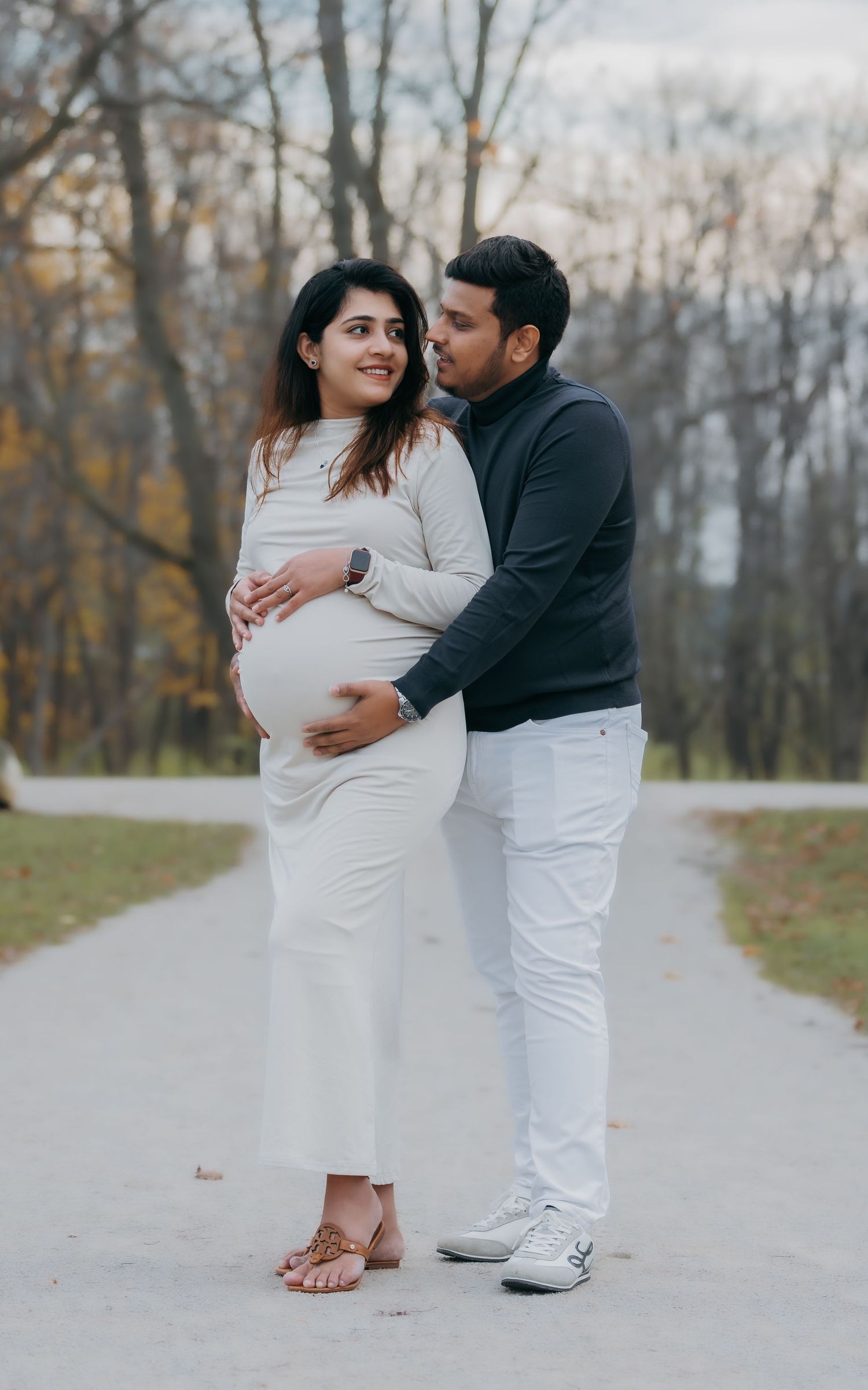 Couple standing on park path, man in navy embracing pregnant woman in white dress from behind.