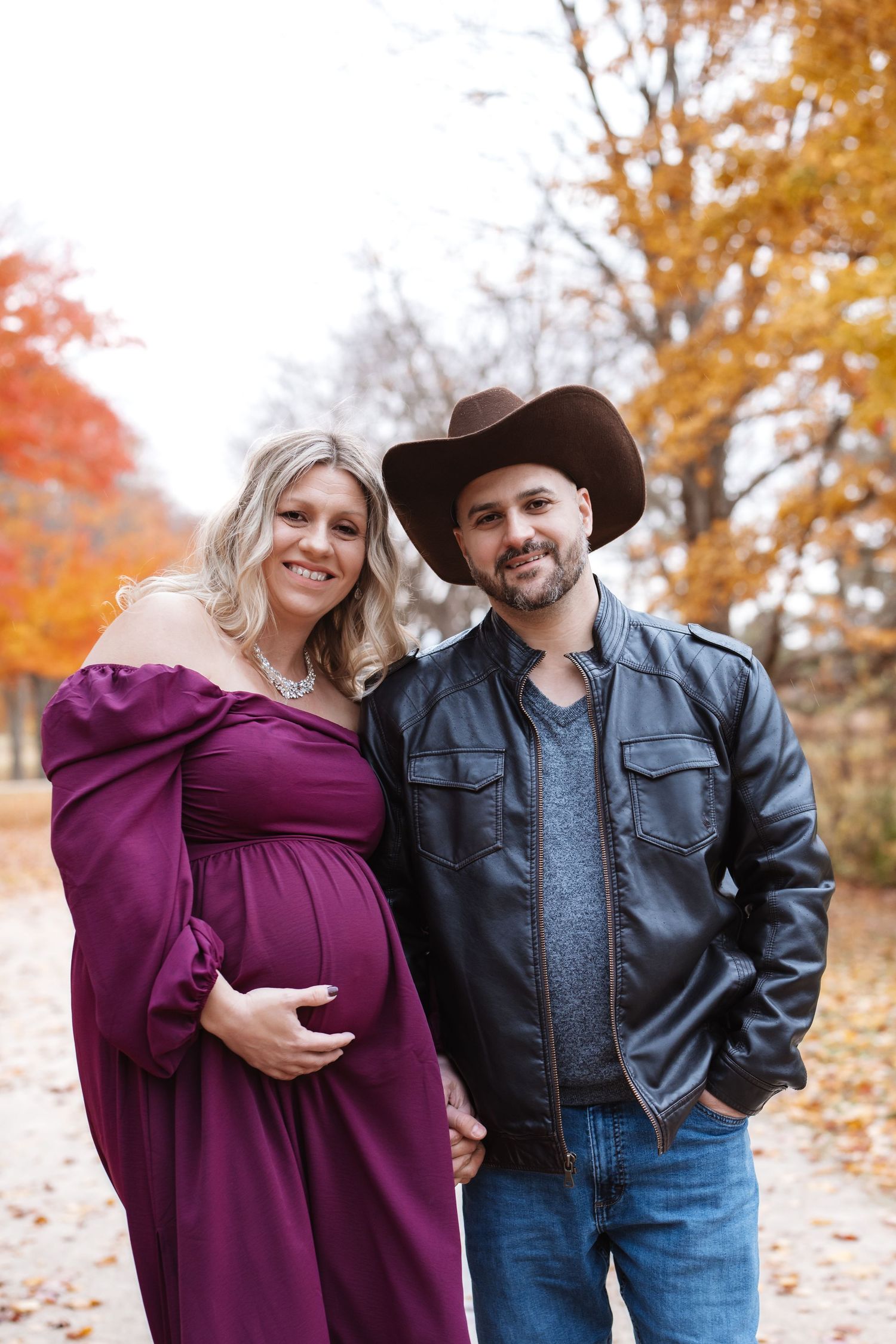 Smiling pregnant couple posing together on an autumn path with colorful fall trees behind them.