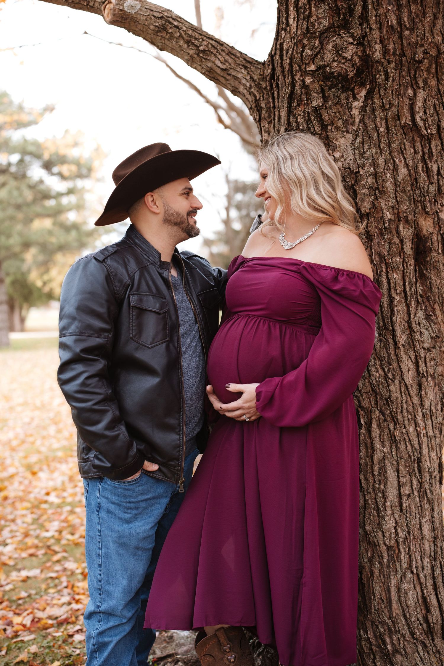 Couple smiling at each other by a tree, man in cowboy hat with pregnant woman in burgundy off-shoulder gown.
