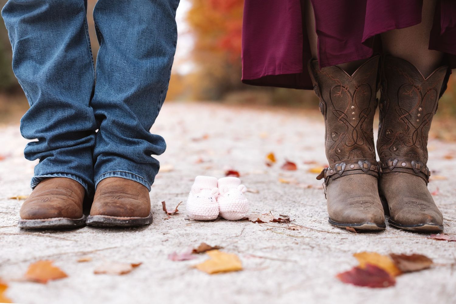 Couple in cowboy boots standing with small pink baby sneakers between them on a leaf-covered path.