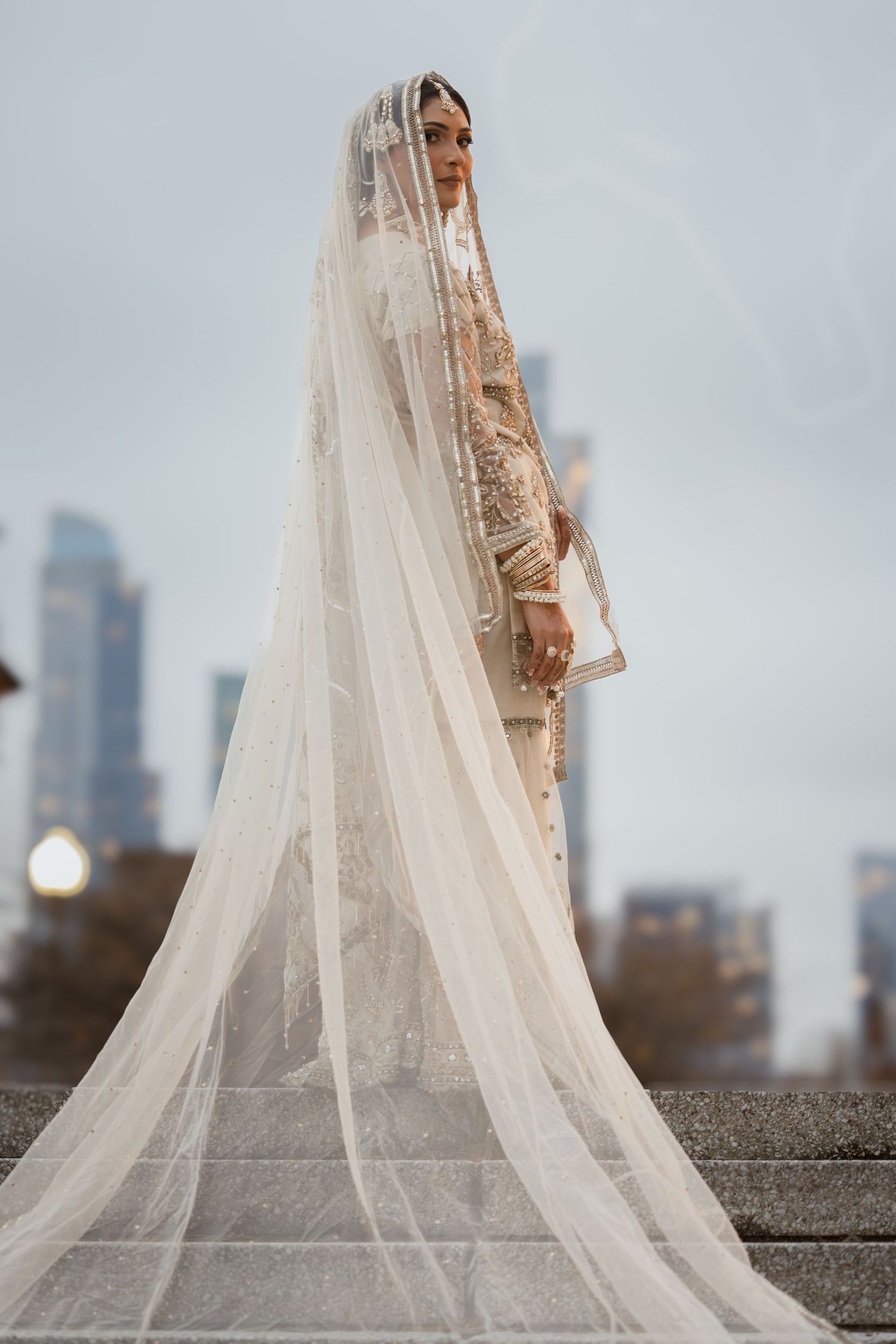 South Asian bride in ivory embroidered lehenga with long cathedral veil on steps with city skyline backdrop.