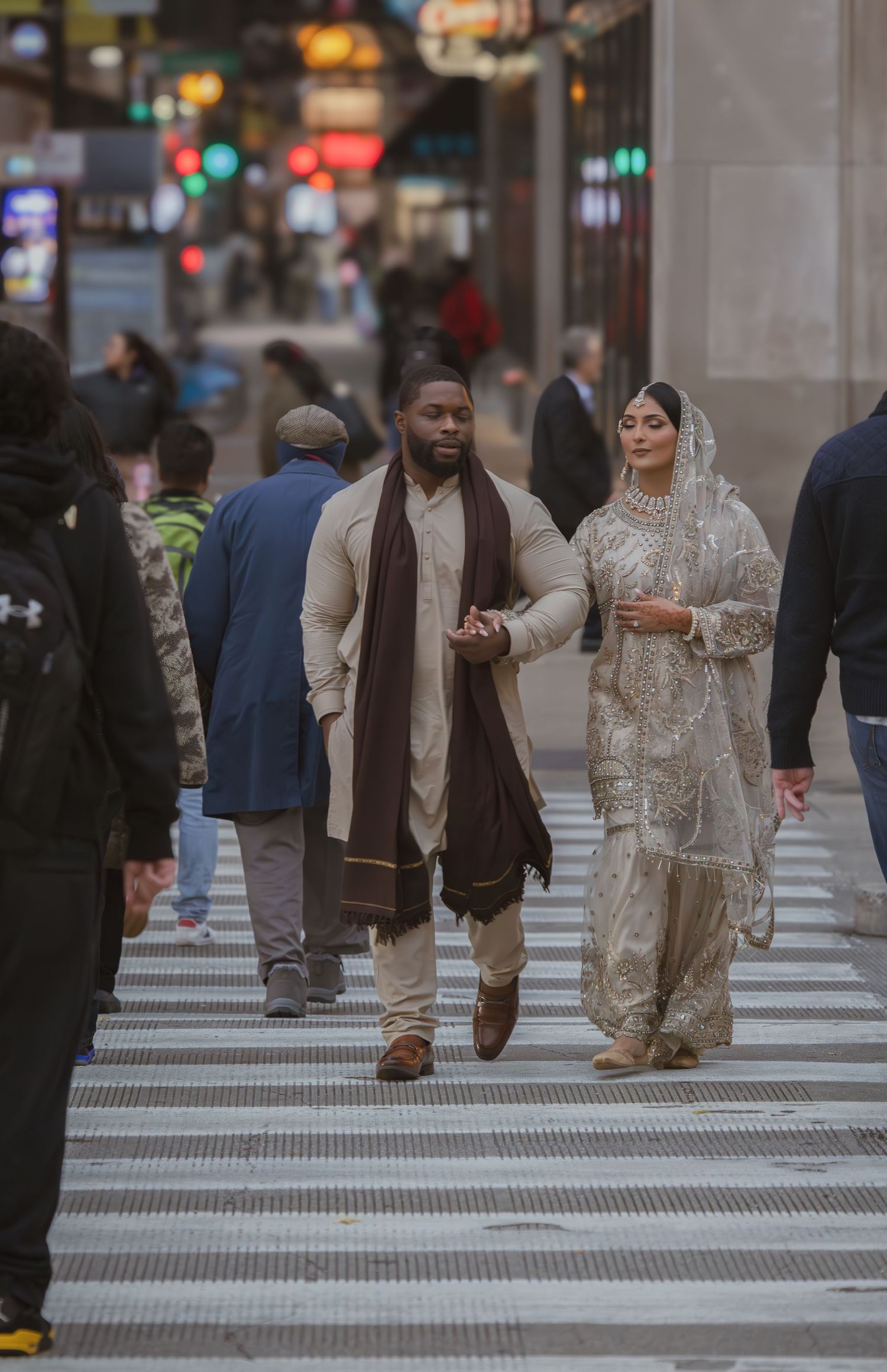 South Asian bride and groom in traditional wedding outfits holding hands while crossing a bustling city street.