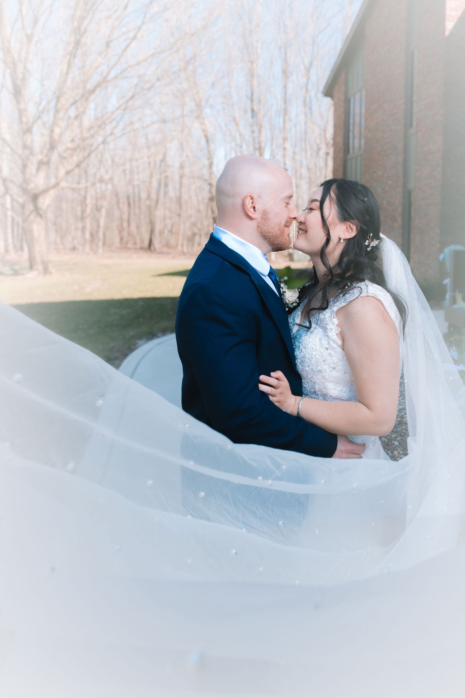 Bride and groom share intimate moment as flowing veil envelops them near brick building.
