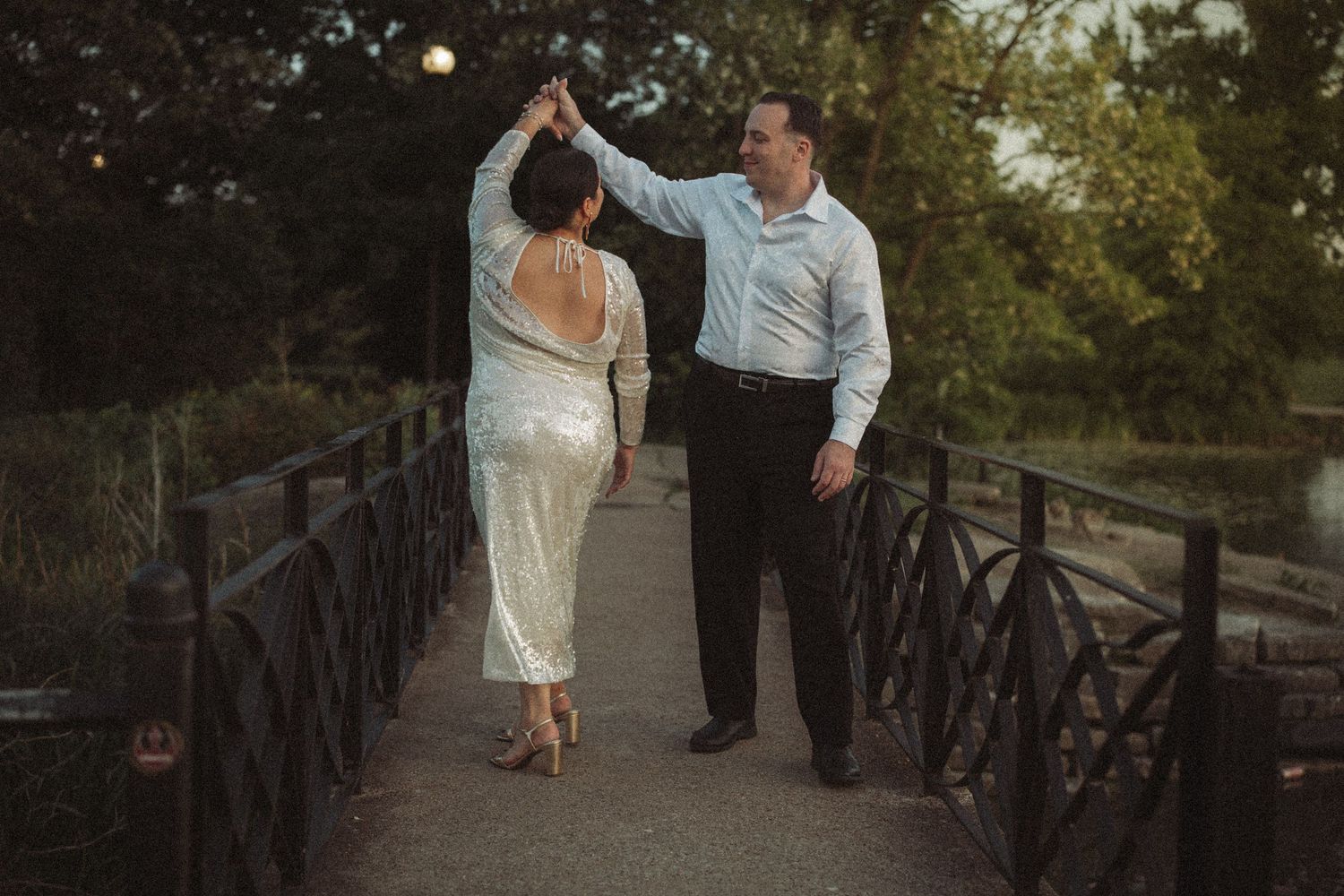 A groom twirls his bride in a sequined dress on a wooden bridge at dusk during their wedding celebration.