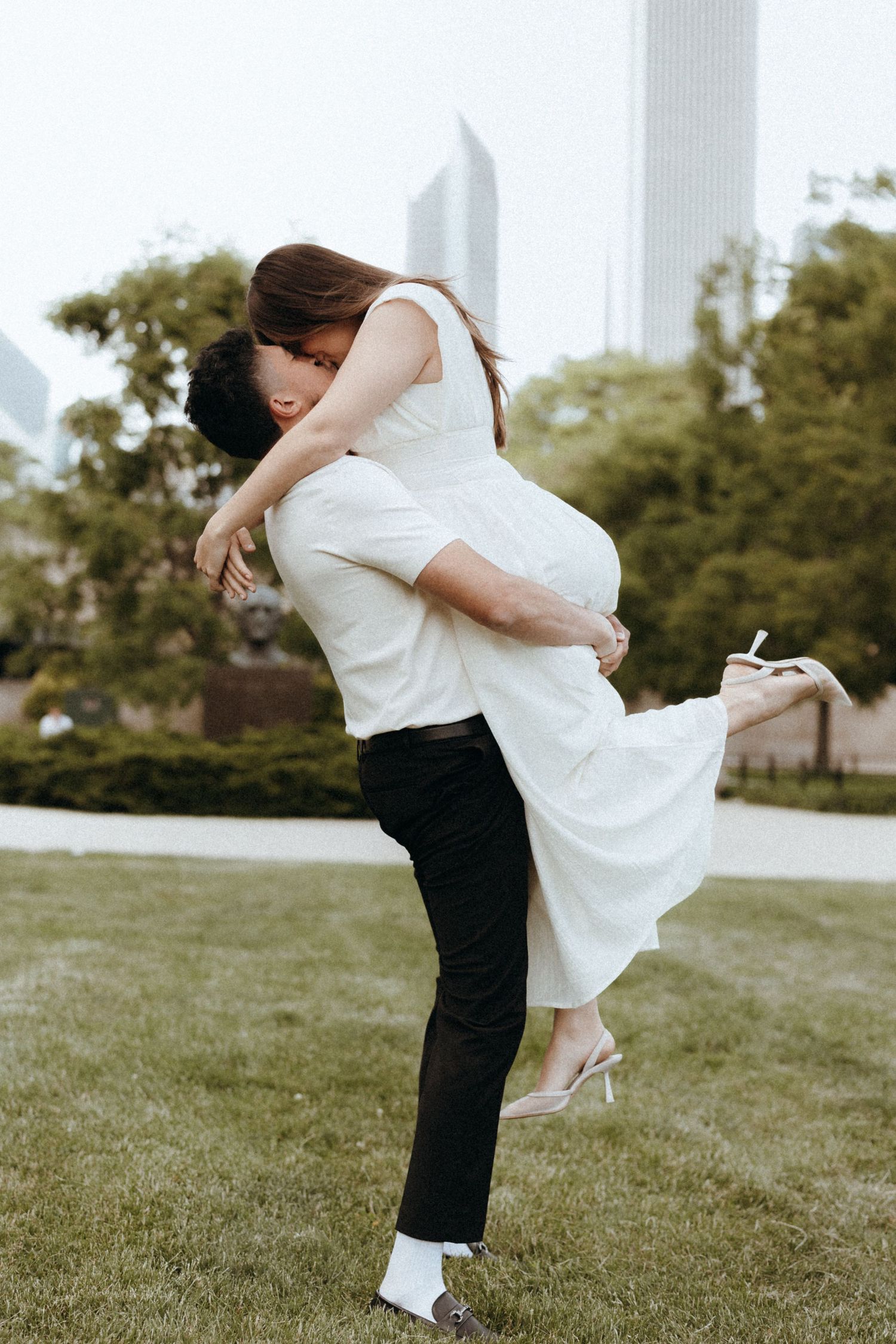 A man lifts and embraces a woman in a white dress outdoors, with city skyline and green trees in the background.