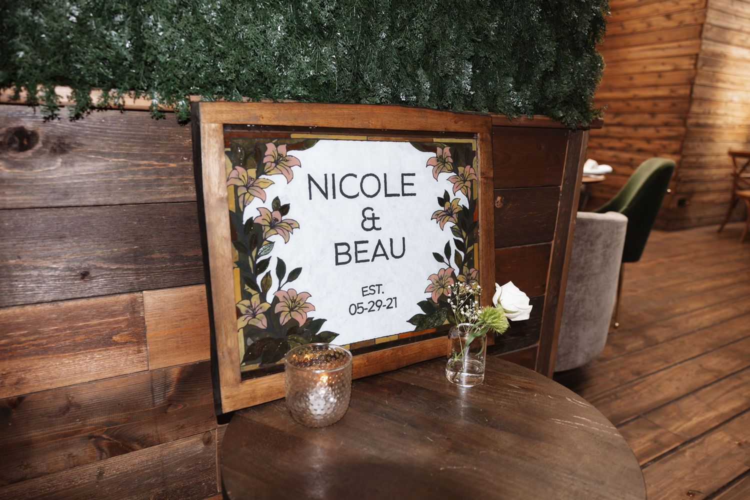 A rustic wooden framed wedding welcome sign reading Nicole and Beau Est. 05.29.21 with floral accents.