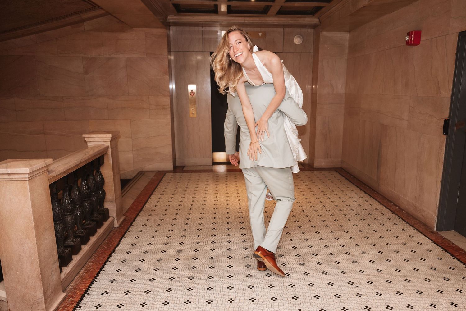 Woman in light gray suit laughing while giving someone a piggyback ride in an elegant hallway with tile floors.