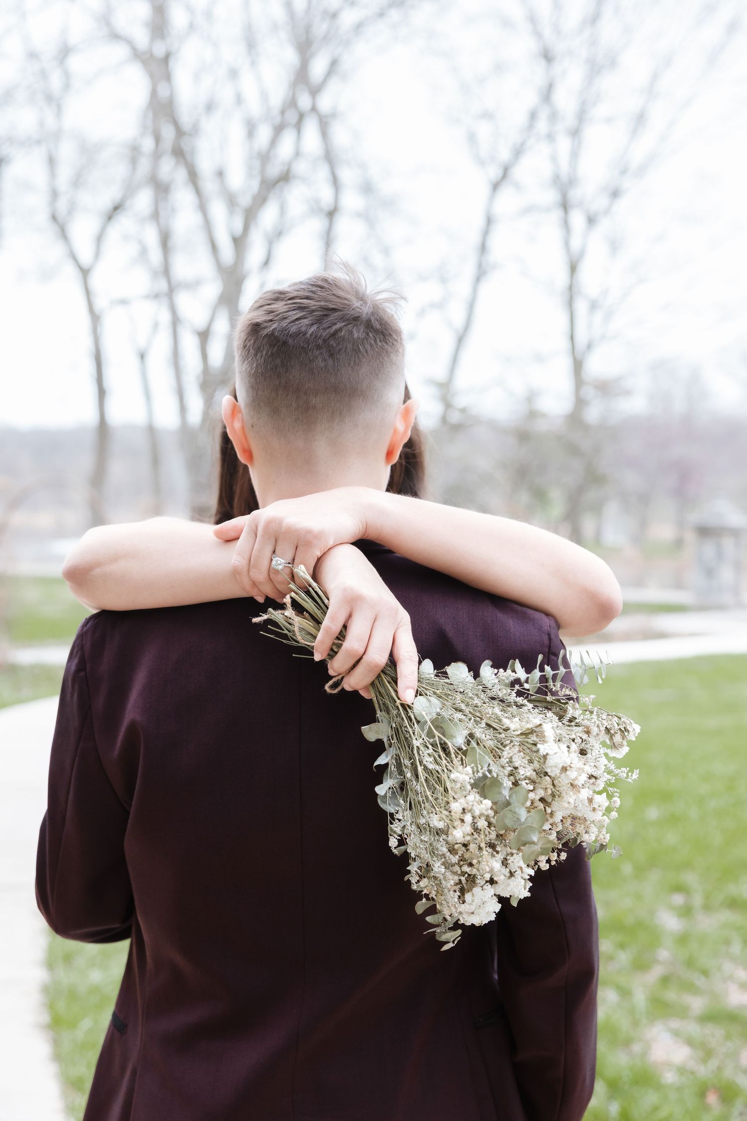 Couple embracing outdoors, woman holding white dried flower bouquet, wearing dark coat, bare trees in background.