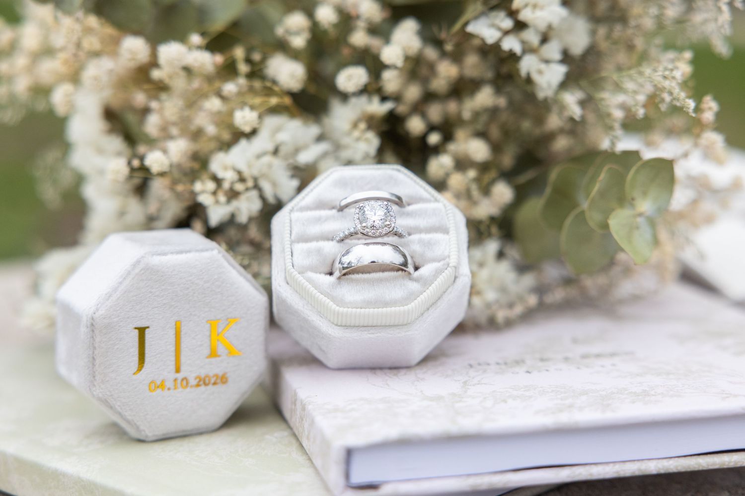 Wedding rings displayed in a monogrammed velvet ring box with J|K initials, surrounded by delicate white flowers and eucalyptus.