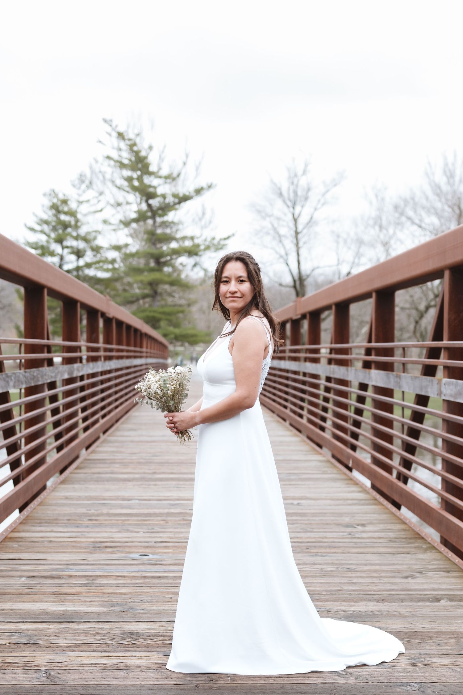 Bride in white gown holding small bouquet stands on wooden bridge surrounded by trees.