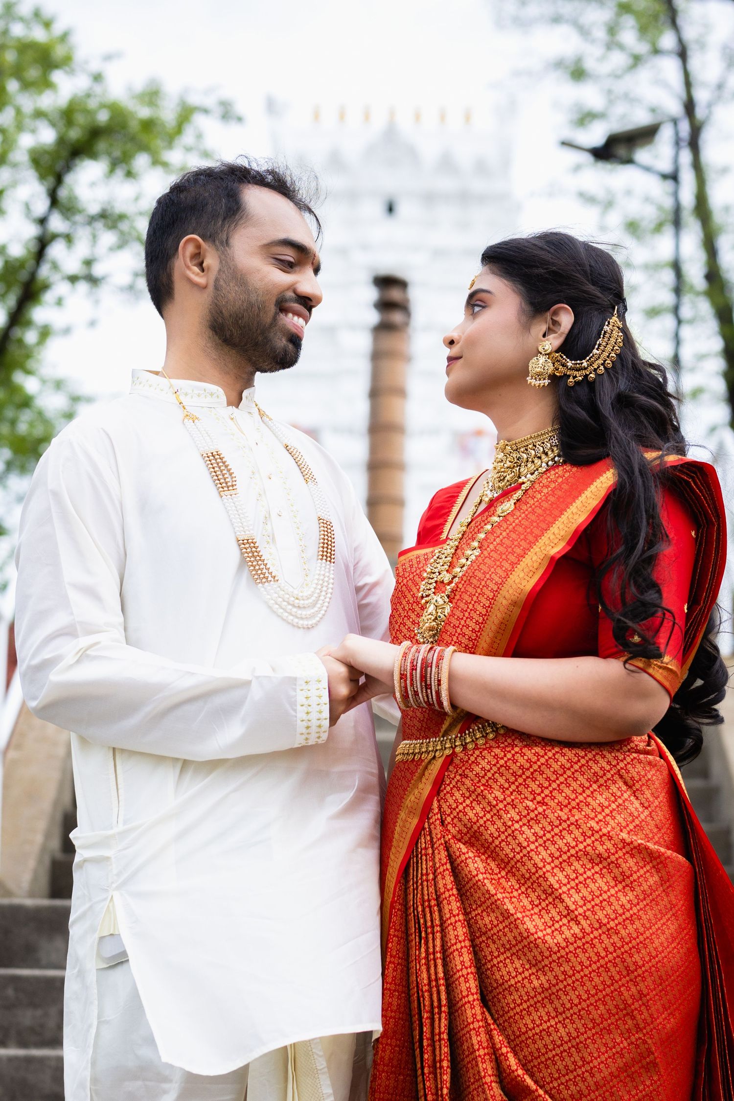 Indian bride and groom in traditional wedding attire holding hands and gazing at each other near white temple.