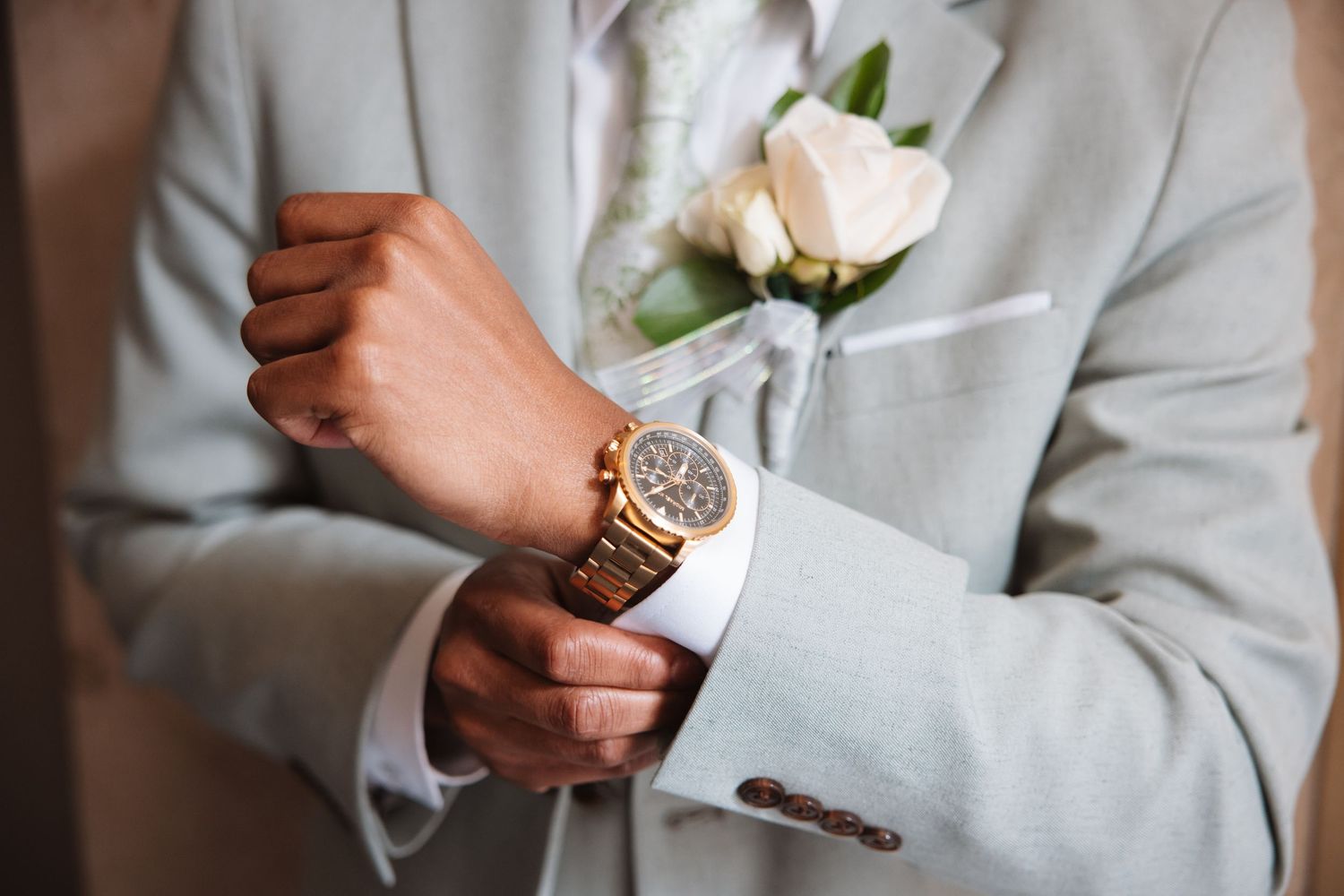 Groom adjusts cufflinks wearing gold watch and light gray suit with white rose boutonniere closeup.