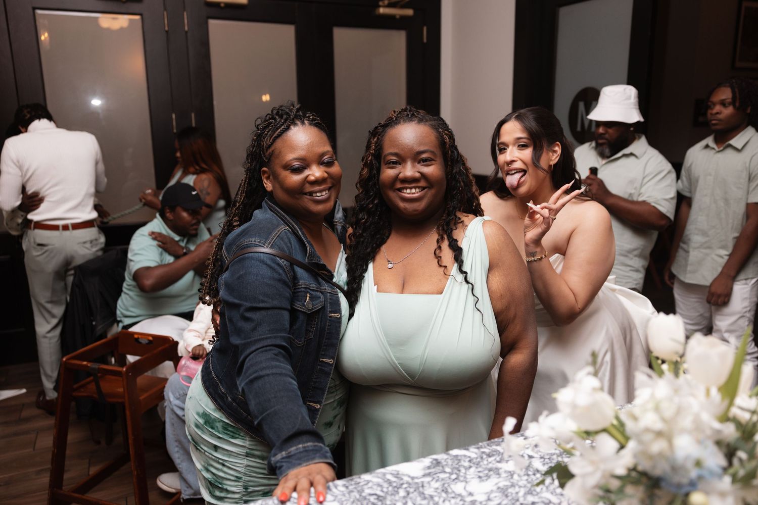 Three smiling women pose together at wedding reception table, one wearing sage bridesmaid dress.