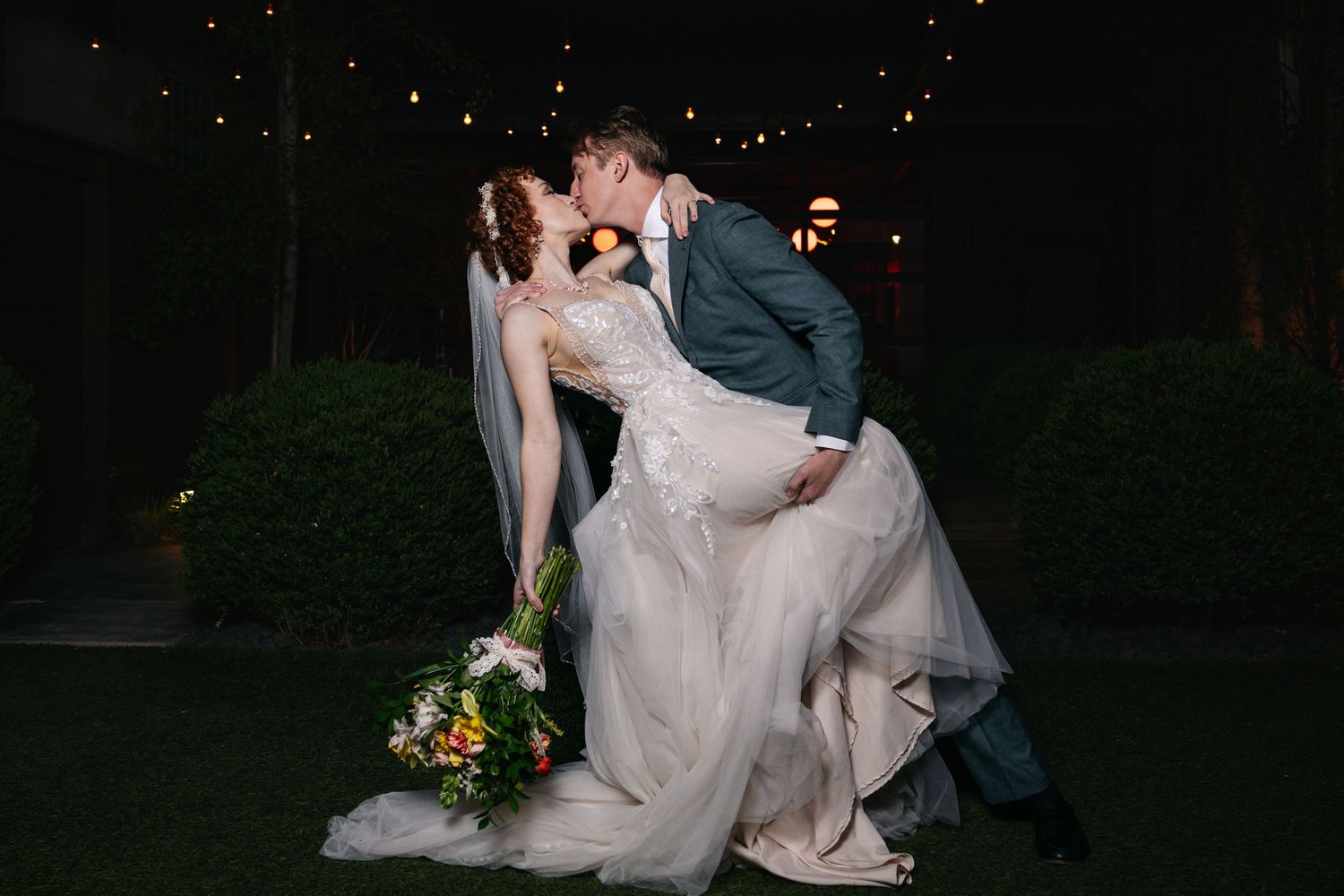 Groom dips bride for a romantic kiss outdoors at night, bride holding a colorful bouquet under string lights.