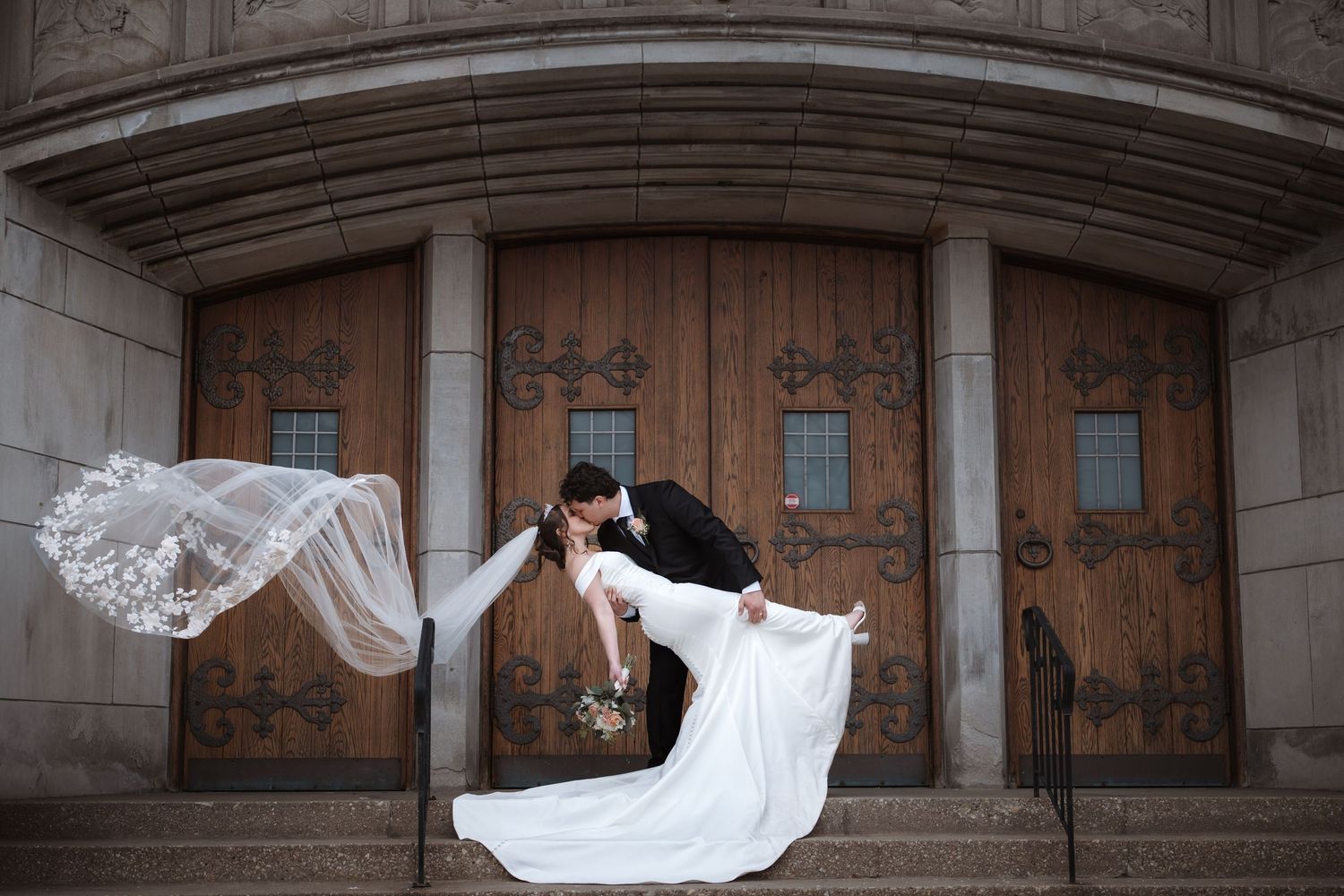 Groom dips bride for a kiss in front of ornate wooden church doors, her lace veil flowing dramatically in the wind.
