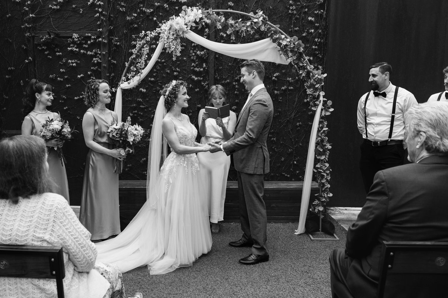 Black and white wedding ceremony photo of bride and groom exchanging vows under floral arch with bridesmaids.