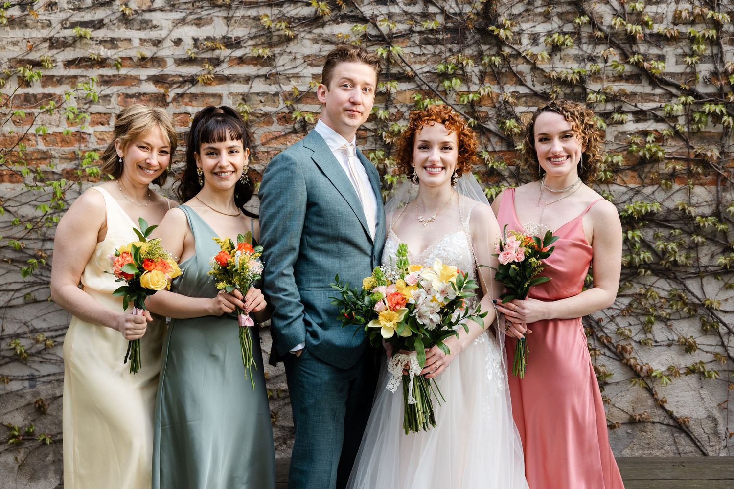 Bride with bridesmaids in colorful dresses holding bouquets of orange and yellow flowers against ivy-covered brick wall.
