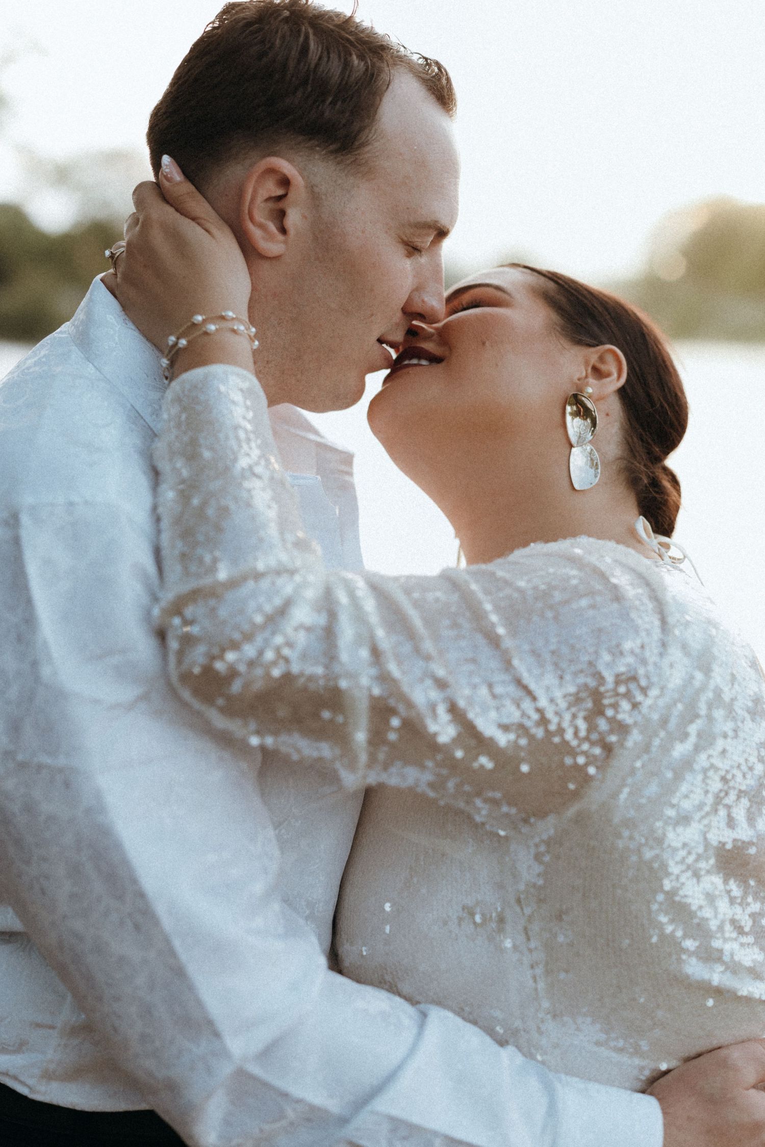 Bride and groom sharing a passionate kiss outdoors in sparkling sequin attire by a sunlit lake.