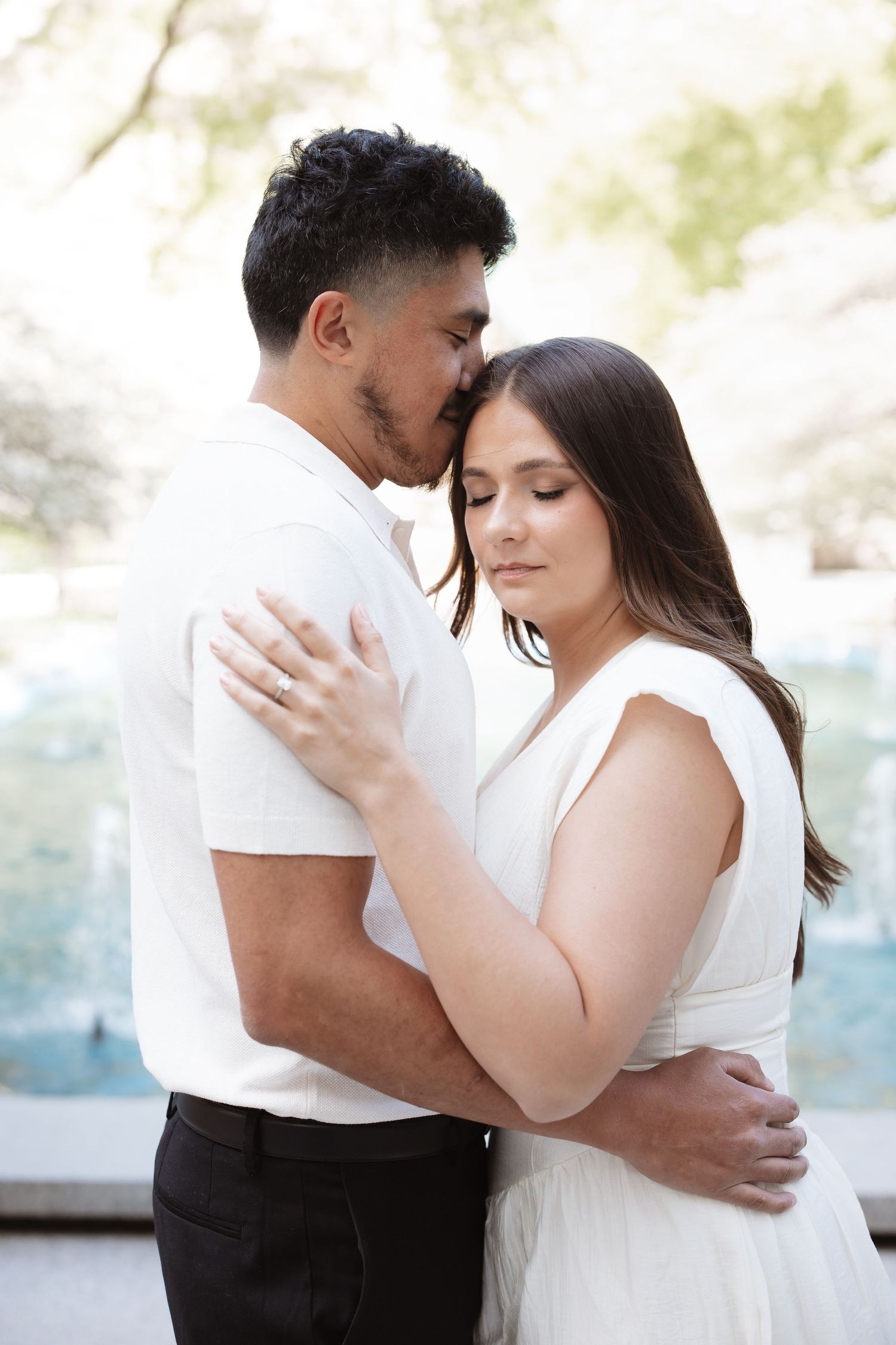 Couple in white embracing tenderly outdoors, woman wearing engagement ring, soft romantic portrait.