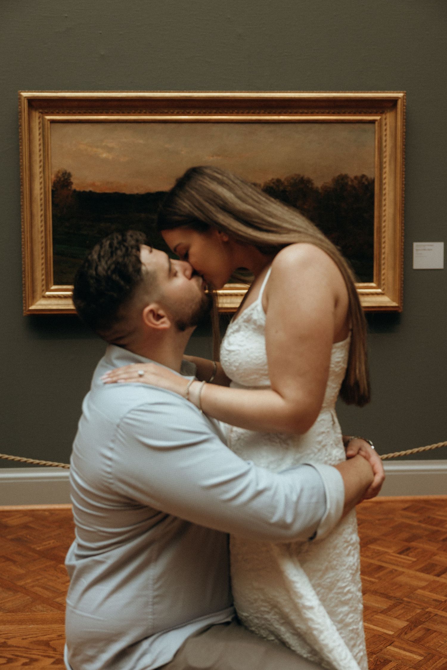 Couple sharing a romantic kiss in front of a gold-framed landscape painting in an art museum gallery.