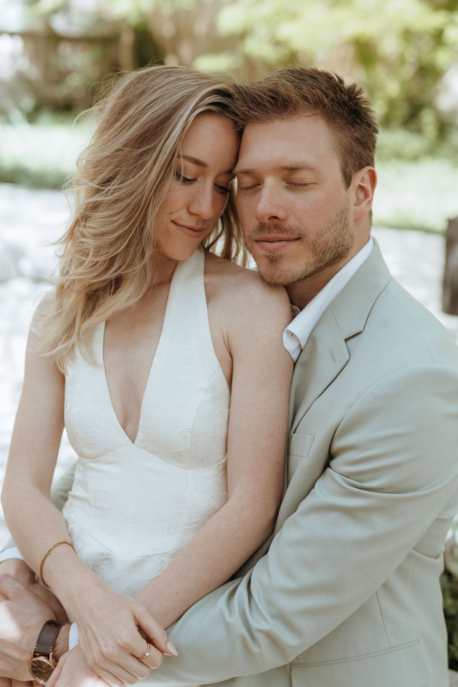 Couple in wedding attire embracing tenderly outdoors, bride in white halter dress and groom in sage suit.