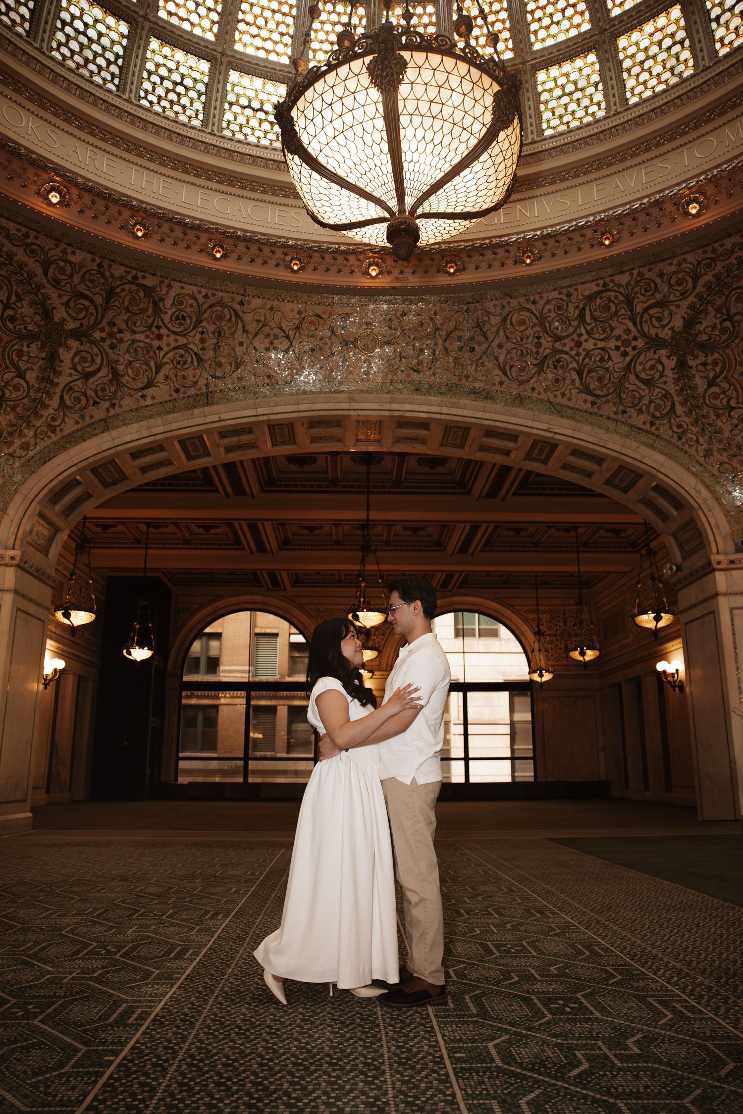 Couple shares romantic embrace under elegant chandelier in ornate ballroom.
