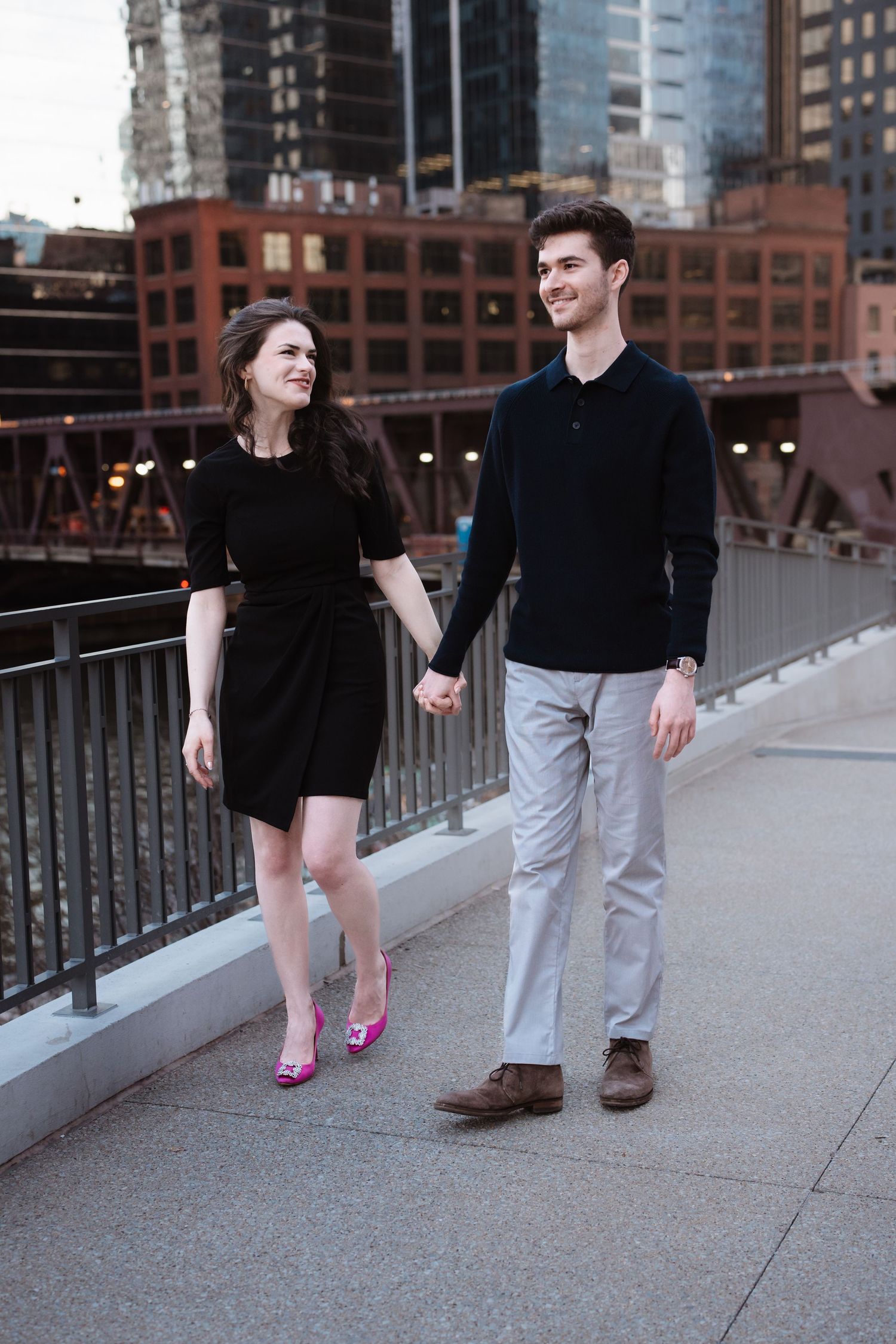 A smiling couple walks hand in hand along a Chicago riverwalk with city buildings behind them.