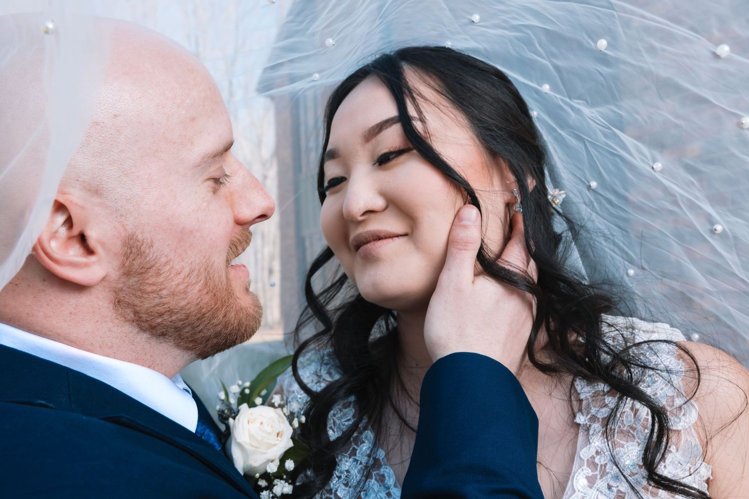 Groom in navy suit gazing lovingly at smiling bride under sheer veil adorned with pearls.
