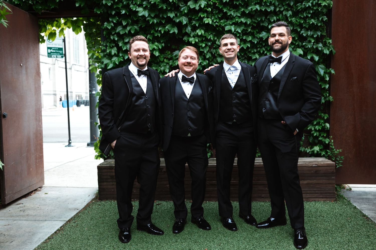 Four groomsmen in black tuxedos with bow ties smiling together outdoors against a green ivy wall.